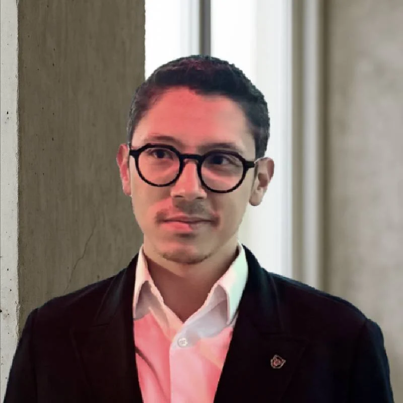 Homem com cabelo castanho, óculos de armação preta, camisa branca e blazer preto, olhando para o lado, na uma sala com parede clara e janela ao fundo.