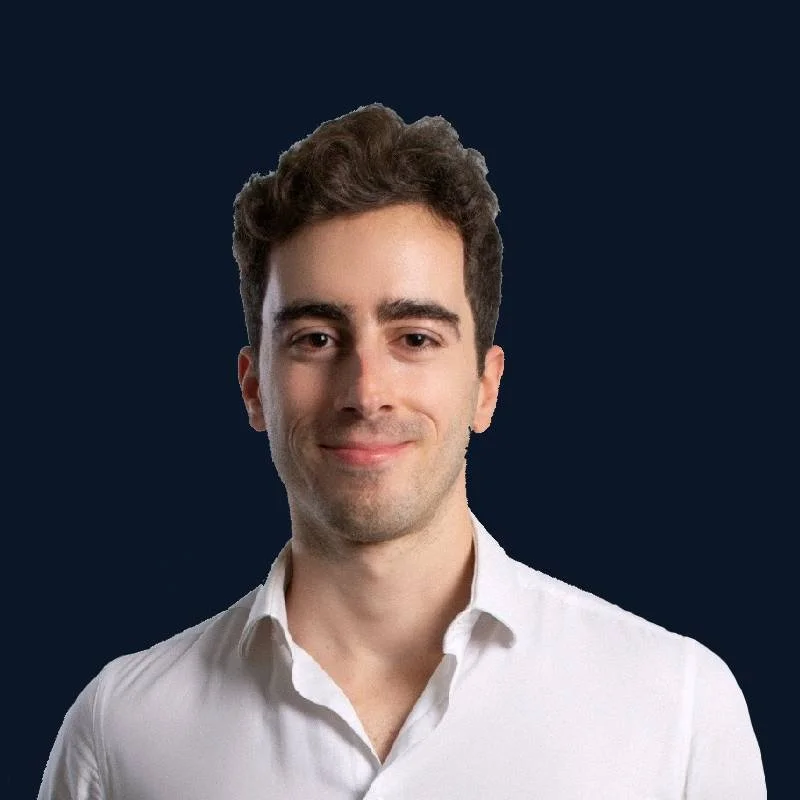 Homem sorridente usando camisa branca, posando ao lado de uma parede de concreto, com janela ao fundo.