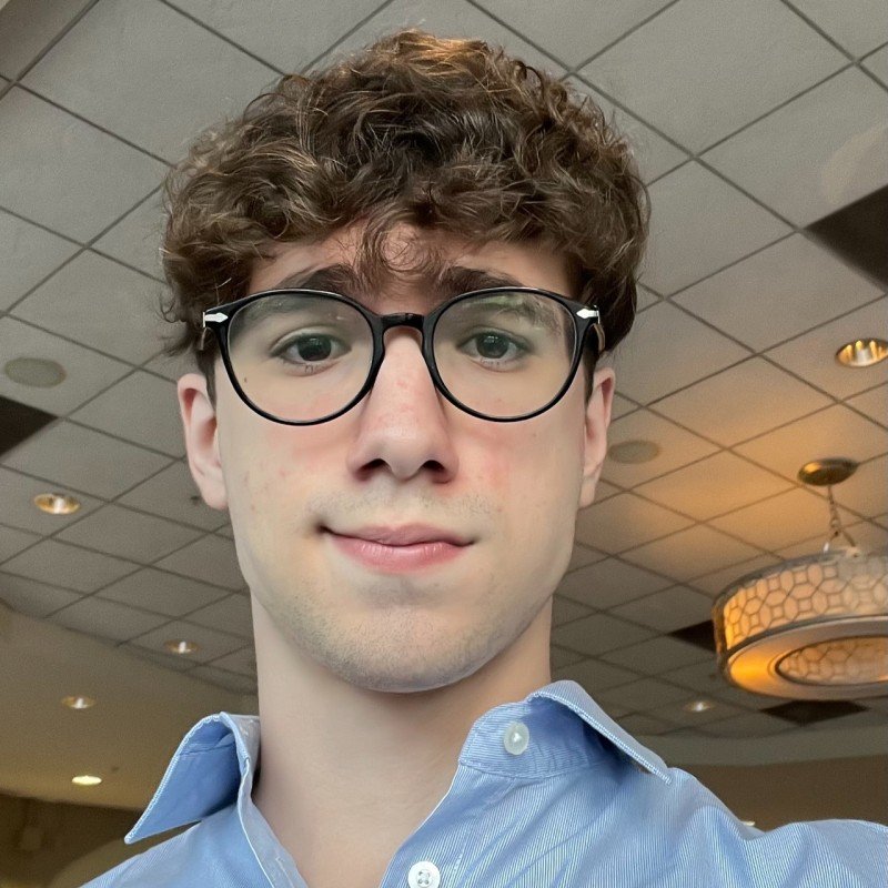 Jovem adulto de cabelo cacheado, usando óculos de armação preta, camiseta azul clara com colarinho e expressão neutra, em ambiente com teto de LEDs e luminária arredondada no fundo.