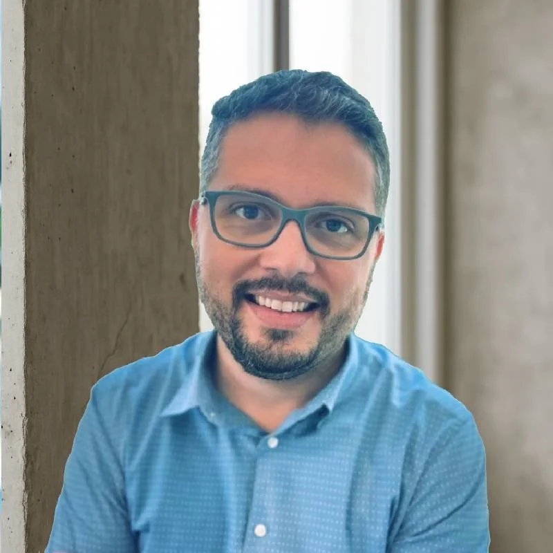 Homem sorridente usando óculos de grau e camisa azul, com fundo de parede e janela.