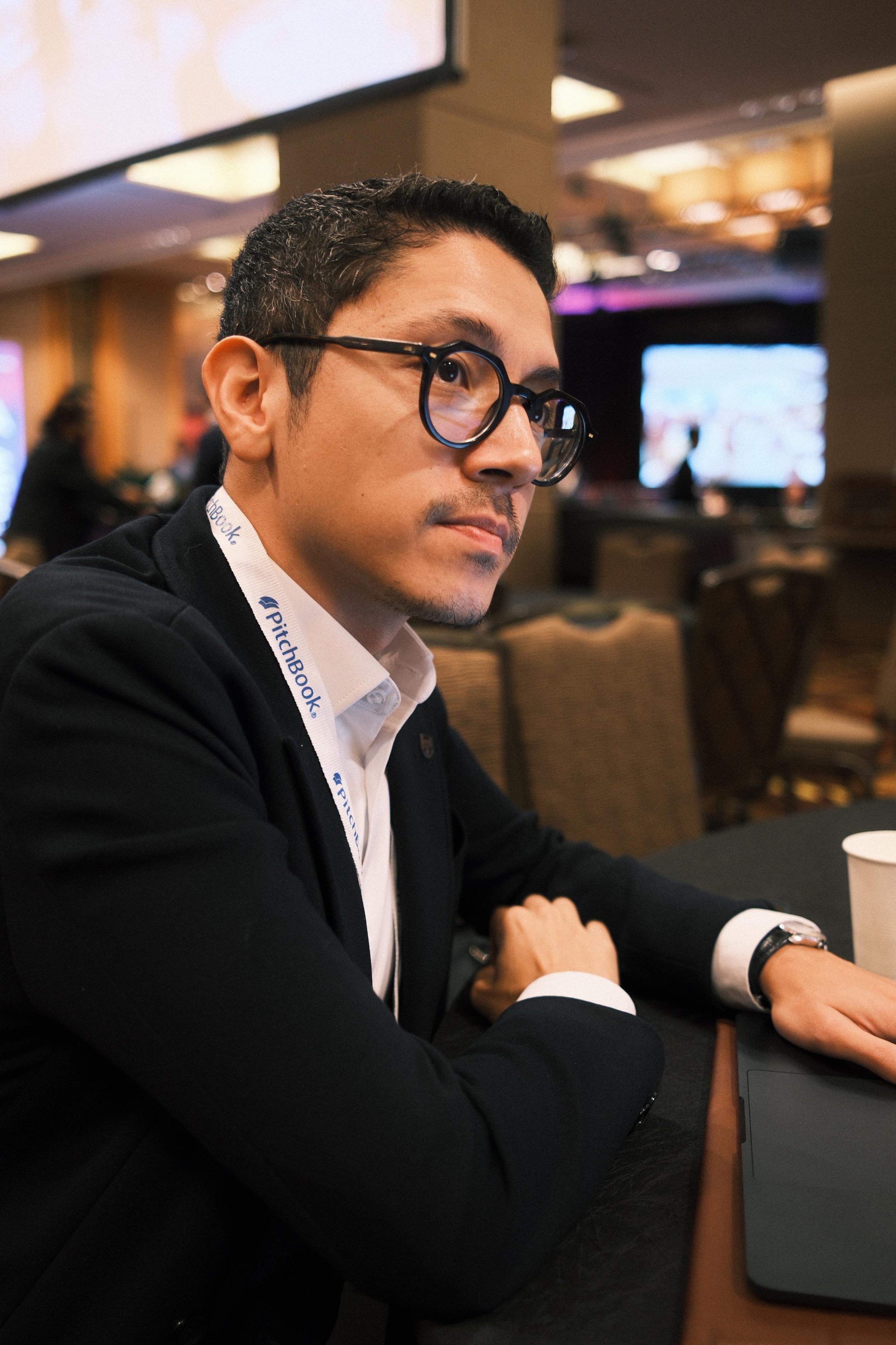 Homem de cabelo escuro, barba por fazer, usando óculos de armação preta, terno preto e camisa branca sentando em uma mesa de evento, com uma barra de acreditação do PitchBook no pescoço, em ambiente de conferência ou reunião.