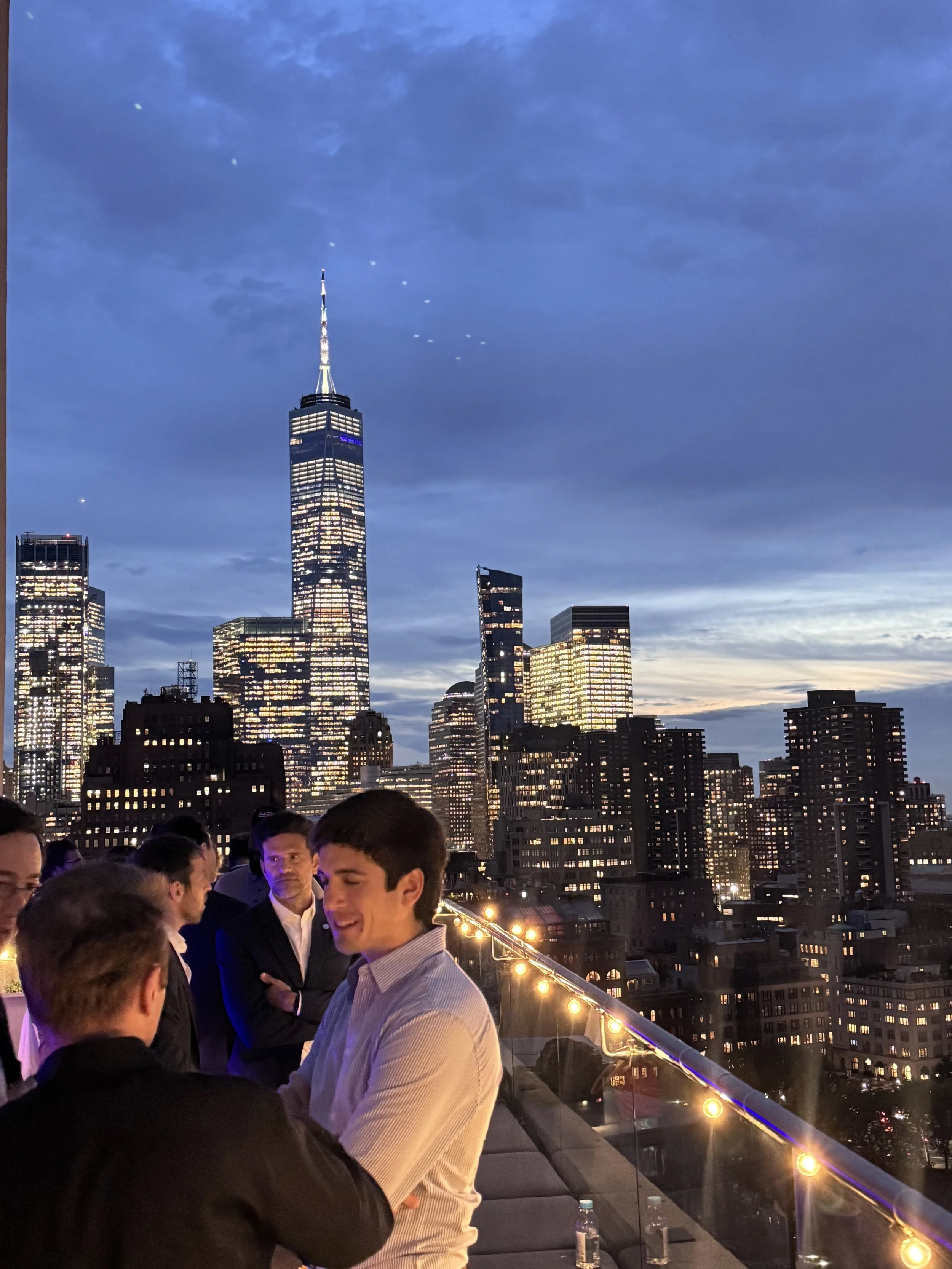 Pessoas conversando em um evento na cobertura de um prédio, com vista para o horizonte de Nova York ao entardecer, incluindo o Empire State Building iluminado.