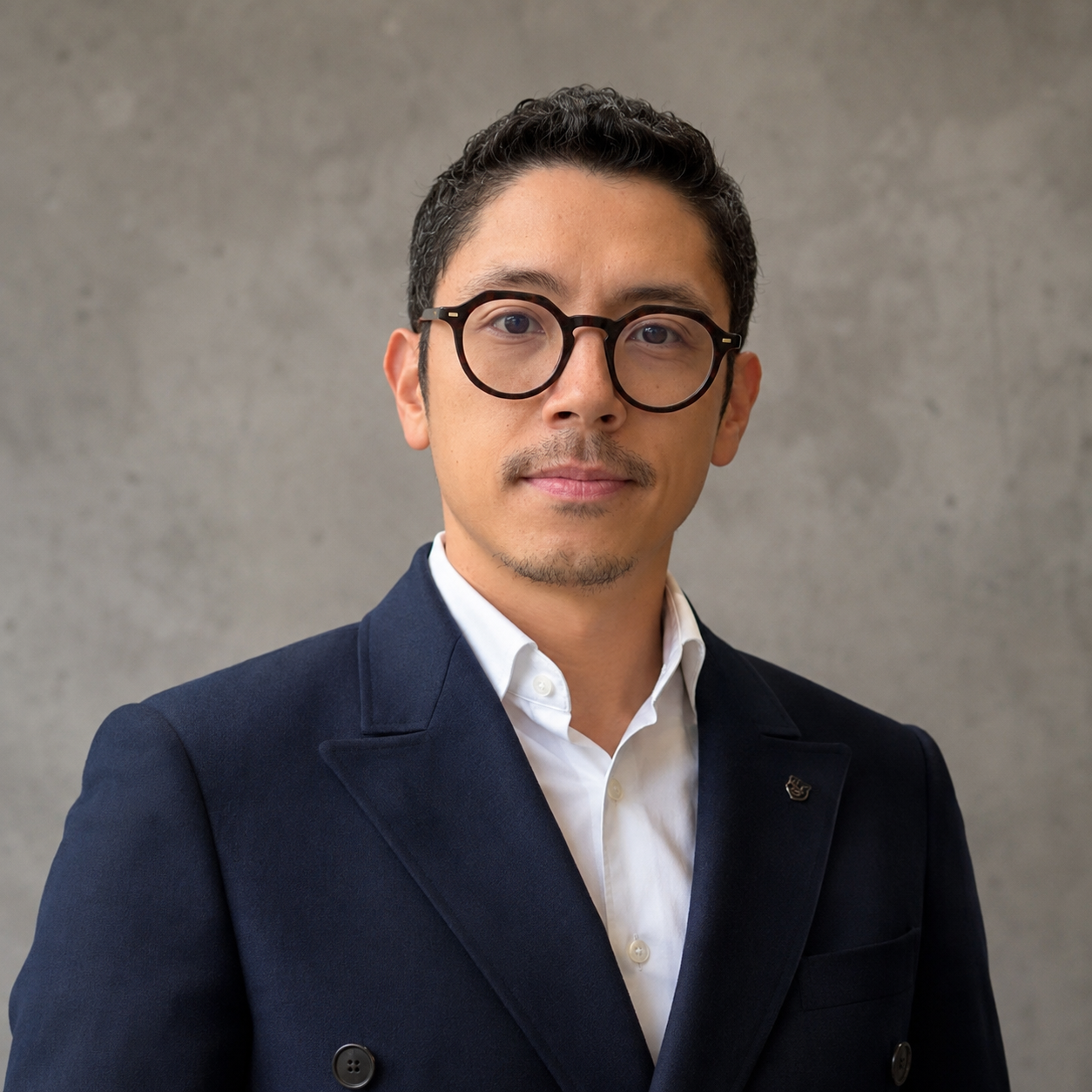 Homem com cabelo castanho, óculos de armação preta, camisa branca e blazer preto, olhando para o lado, na uma sala com parede clara e janela ao fundo.