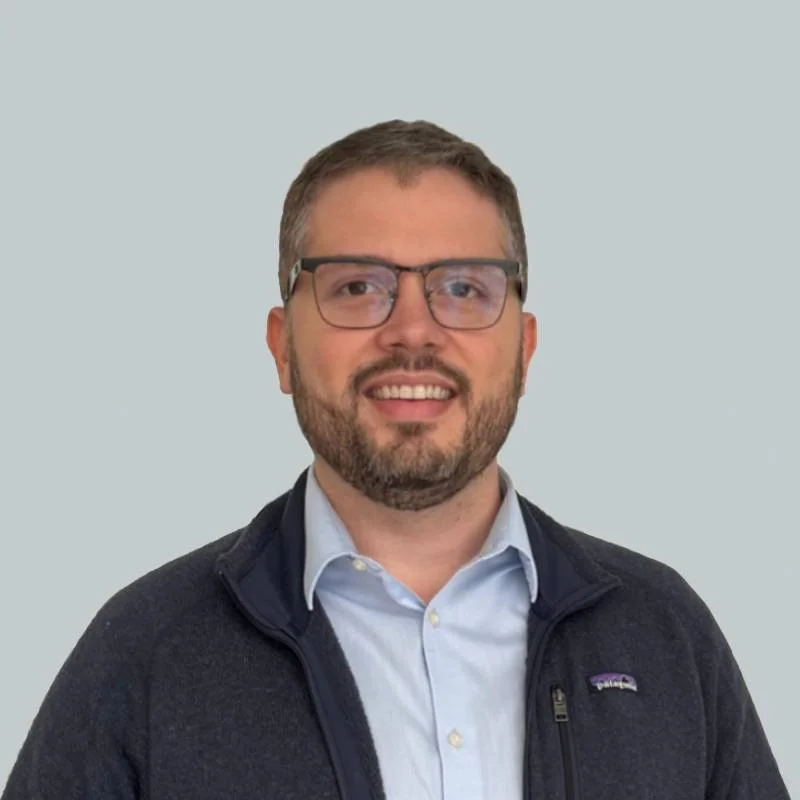 Homem sorridente usando óculos de grau e camisa azul, com fundo de parede e janela.