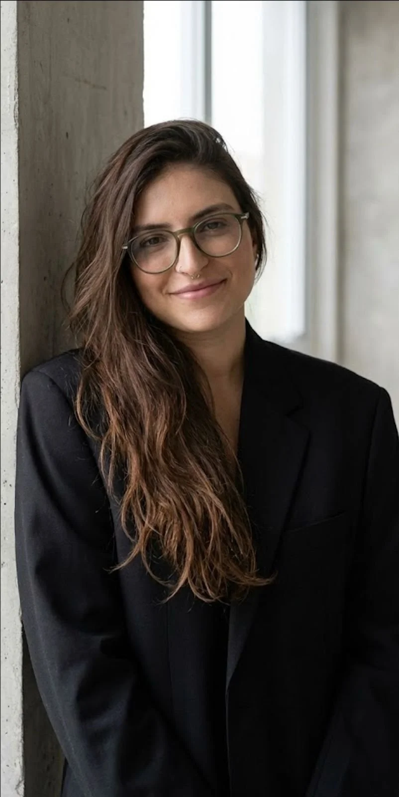 Mulher com cabelo castanho ondulado, óculos de grau e sorriso, usando blazer preto, posando junto a uma parede de cimento em ambiente de iluminação natural.