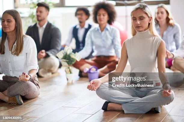 A group of diverse people participating in a meditation session in a bright room, sitting cross-legged with eyes closed.