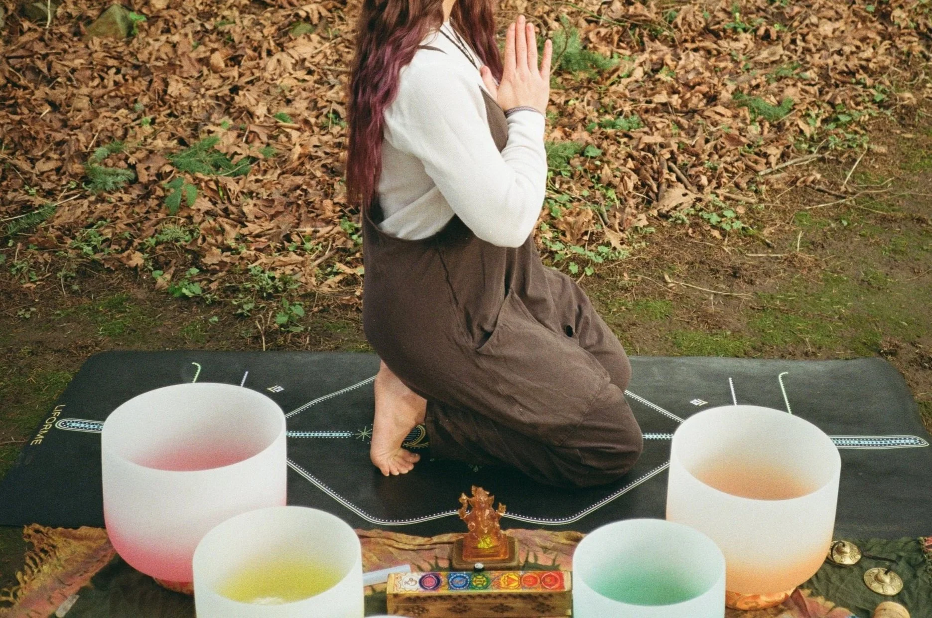 A woman kneeling outdoors on a black mat with her hands in a prayer position, surrounded by singing bowls and spiritual objects, with a natural background of leaves and grass.