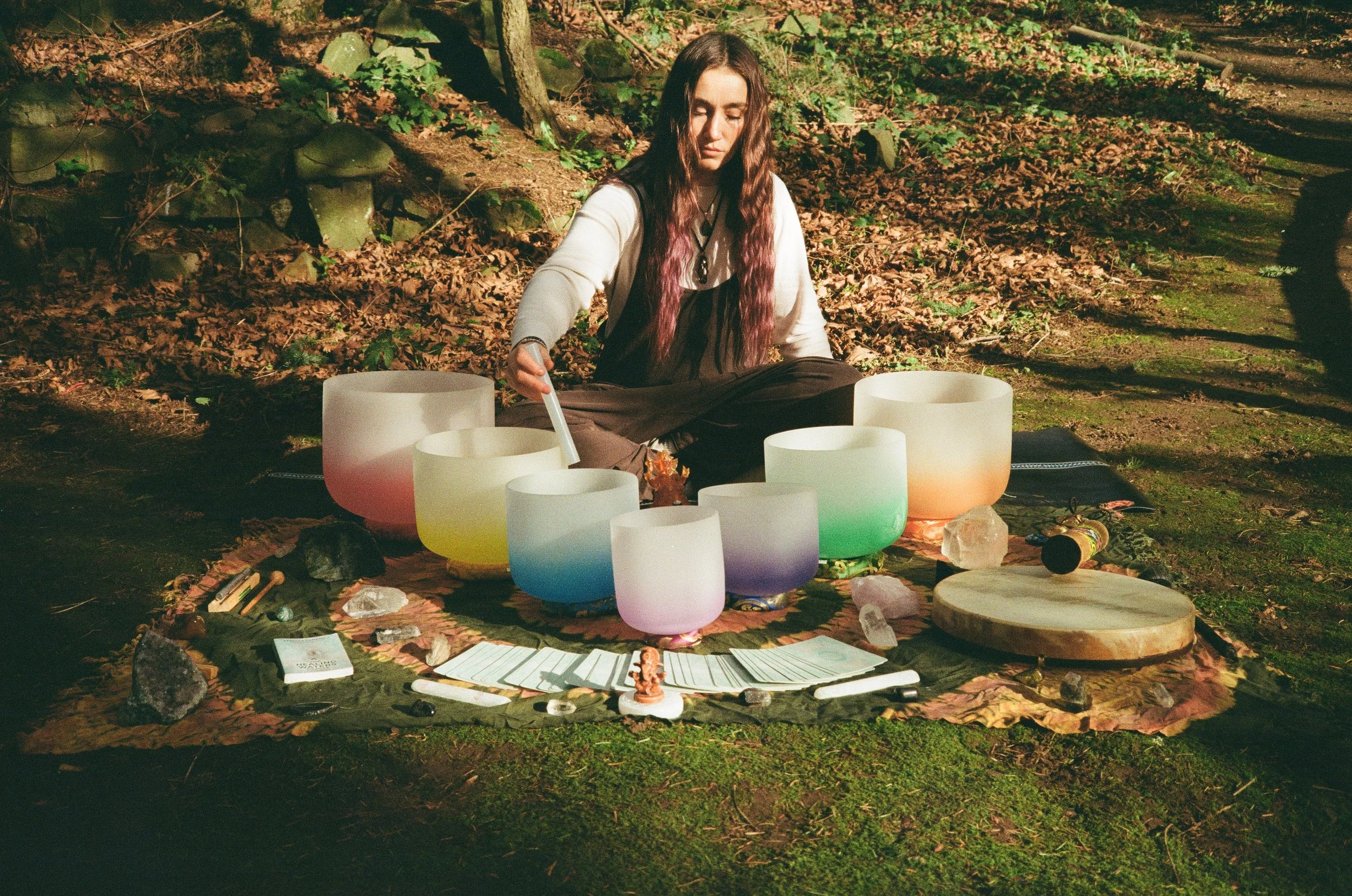 A woman with long brown hair sits cross-legged outdoors on the ground, playing crystal singing bowls surrounded by various crystals, a wooden board, and sheets of paper, with fallen leaves and trees in the background.