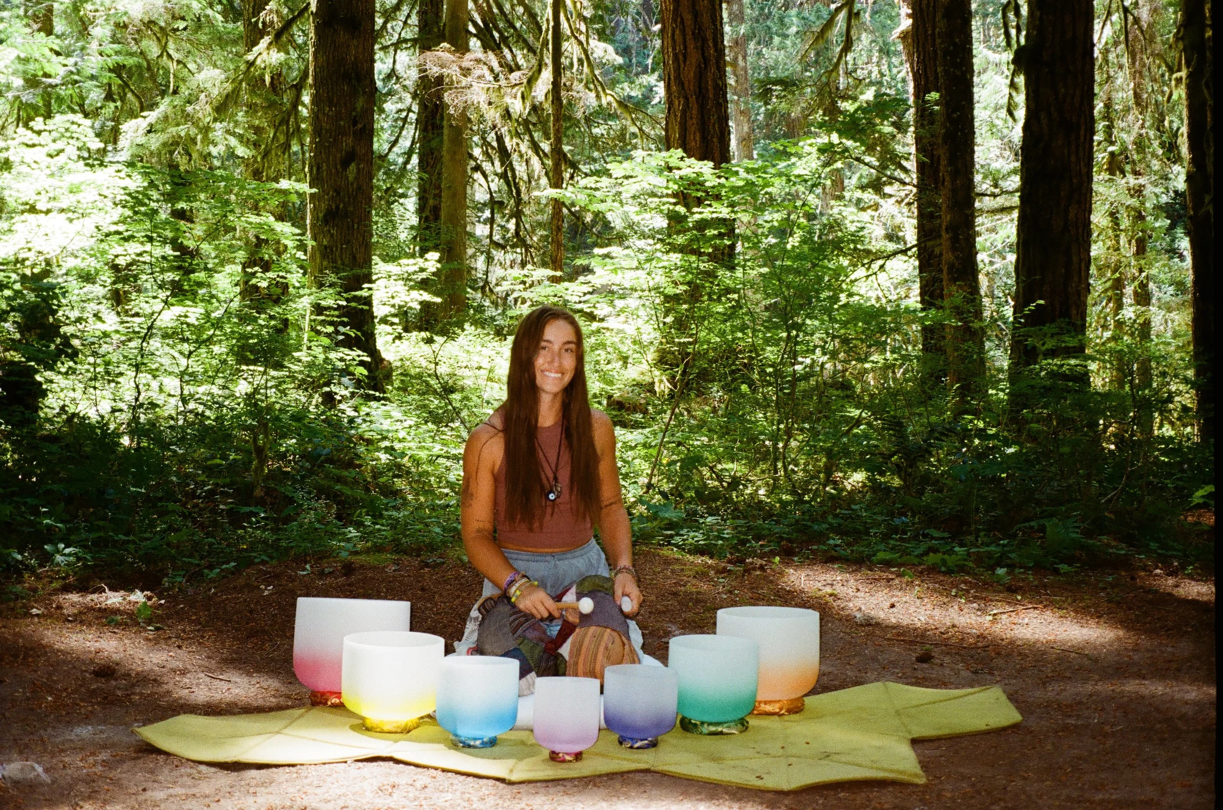 A young woman sits in a forest with a collection of illuminated singing bowls placed on a yellow mat in front of her. She is smiling, wearing a sleeveless top and bracelets, surrounded by tall trees and lush green foliage.
