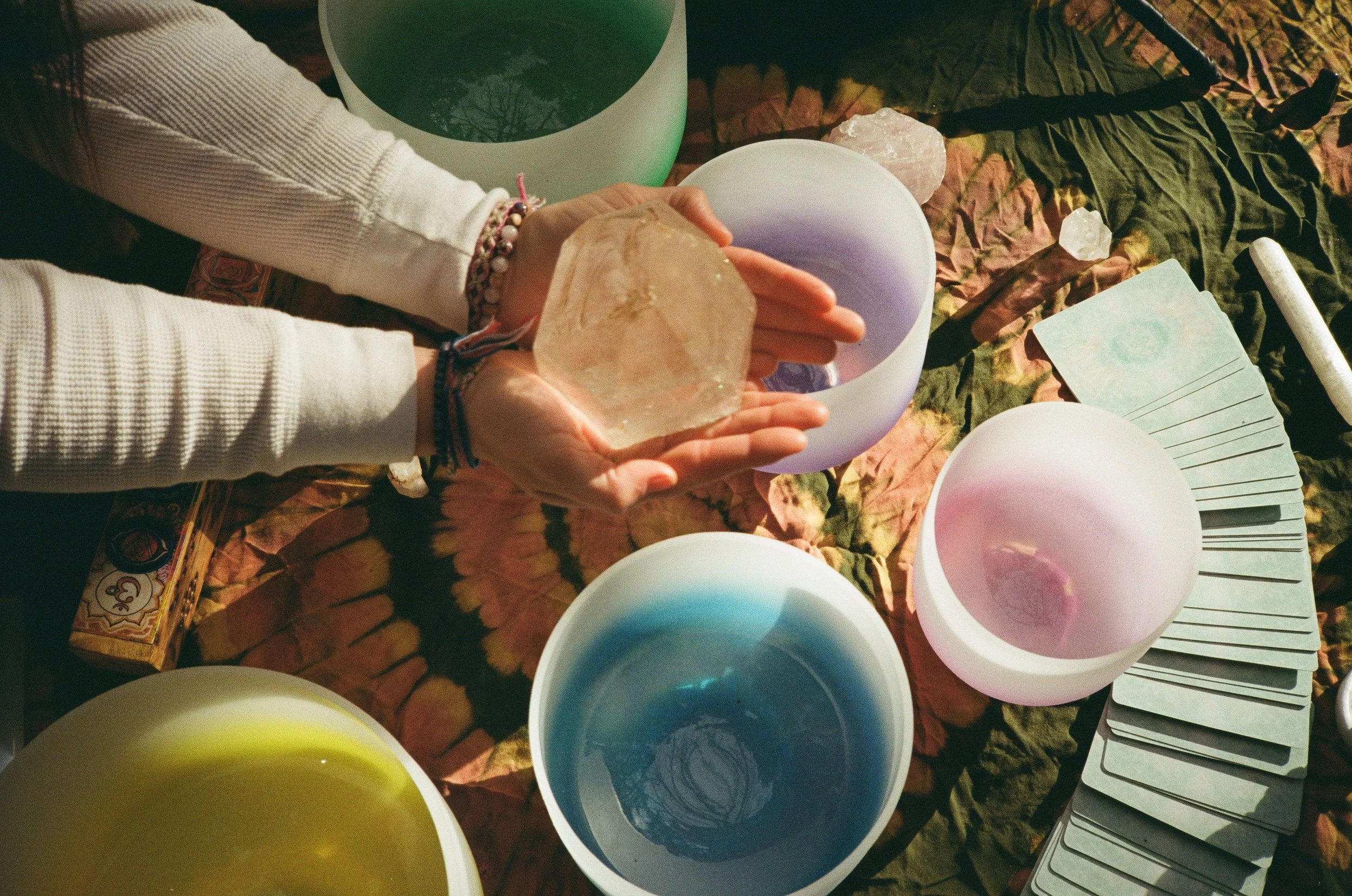 Person holding a large clear crystal above singing bowls with various colors, tarot cards, and a deck of playing cards on a cloth-covered surface.