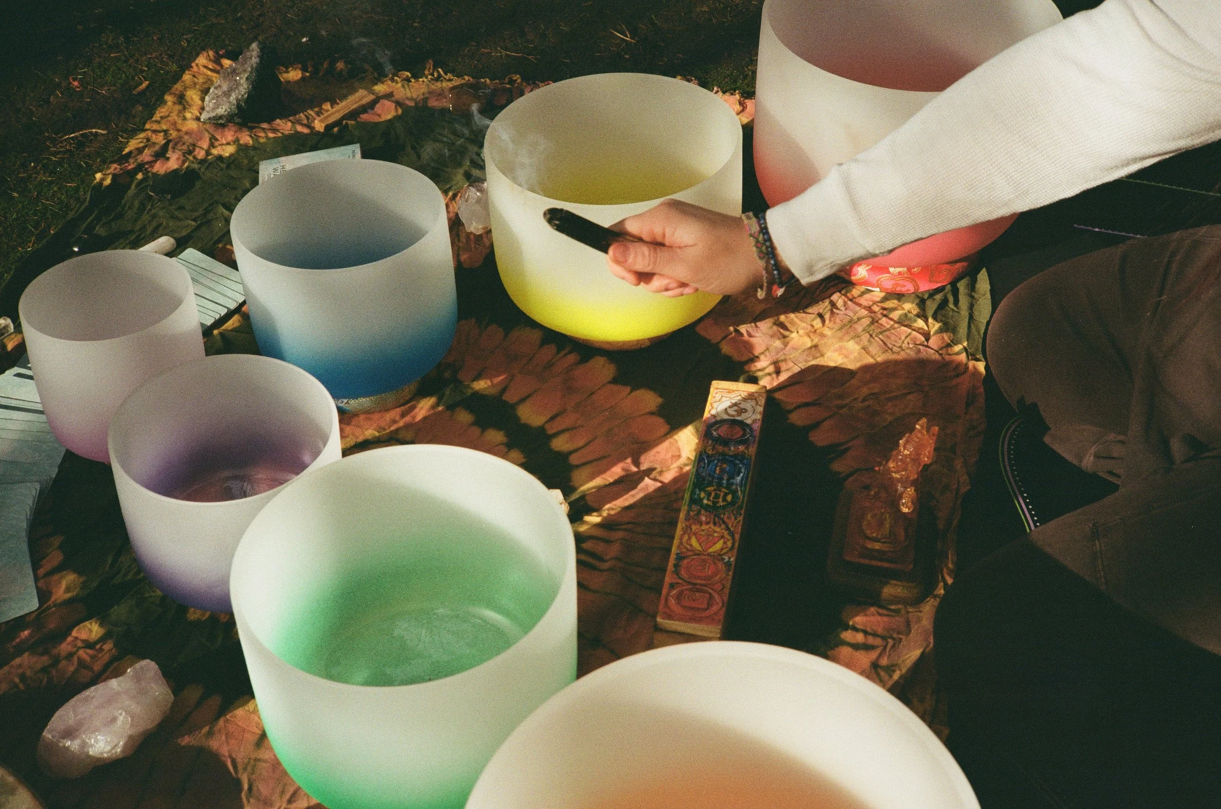 A person playing crystal singing bowls on a patterned cloth, with various colored bowls, tarot cards, and crystals around.