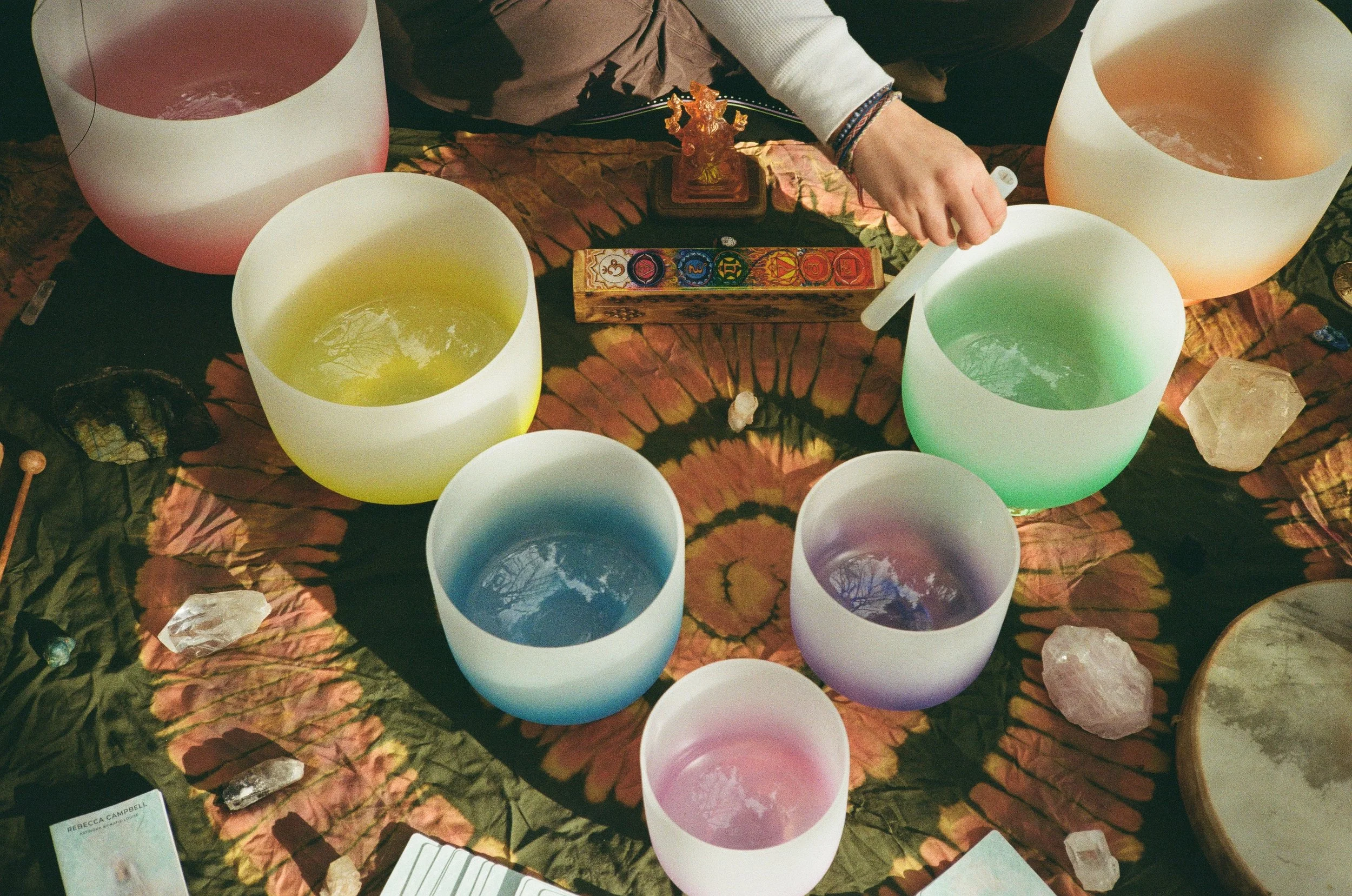 Six large and small singing bowls filled with colored water, arranged in a circle on a tie-dye cloth, with crystals, cards, and a figurine nearby.