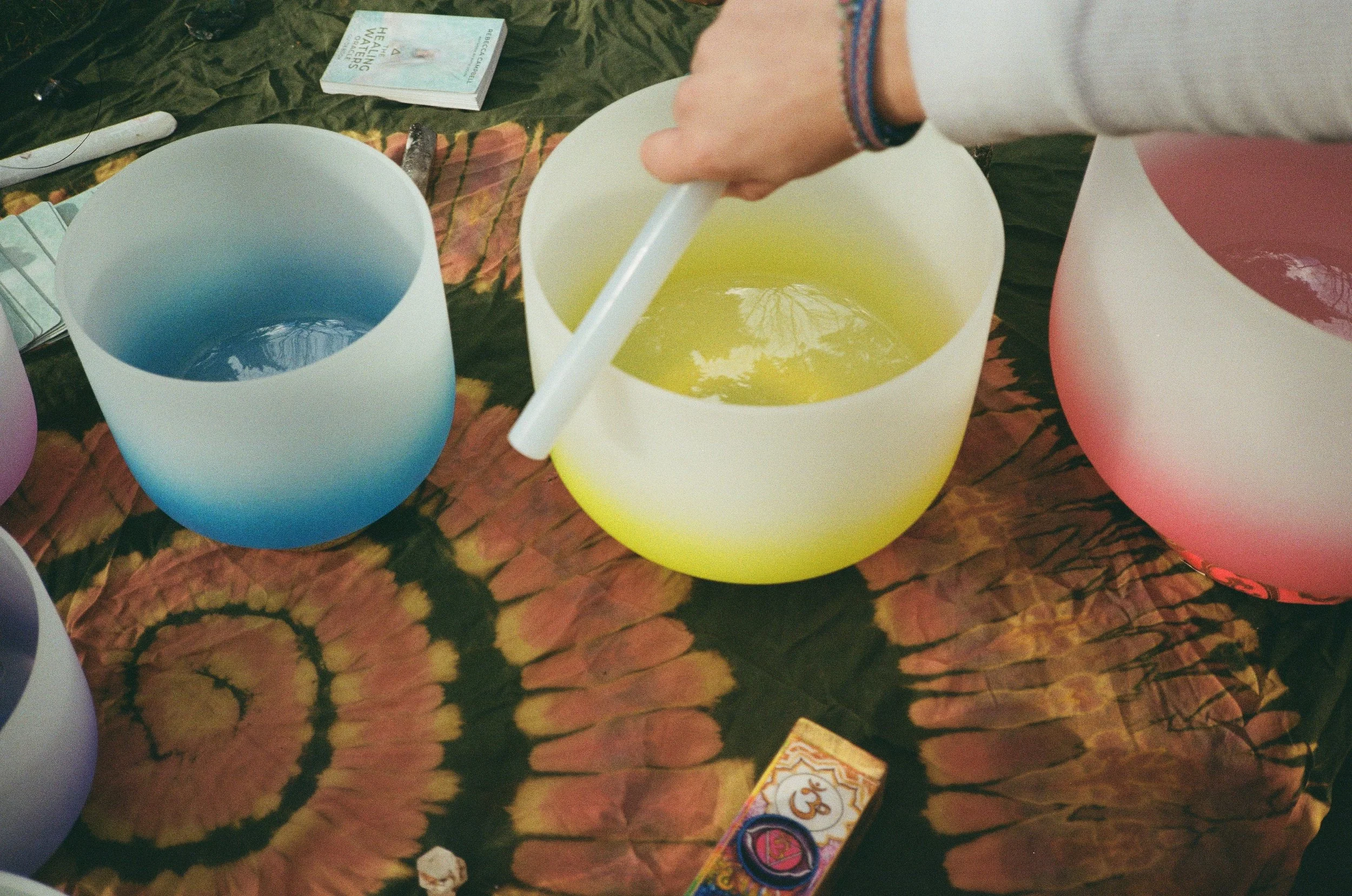 Colorful crystal singing bowls on a tie-dye cloth, with a person stirring the yellow bowl and other bowls nearby, and incense and spiritual cards in the background.