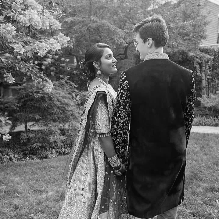 A man and woman dressed in traditional South Asian attire standing outdoors in a garden, facing each other and holding hands.