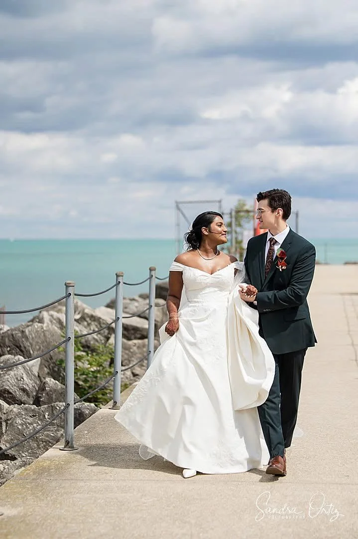 A Chicago bride in a white wedding dress walking alongside a groom in a black suit along a Lake Michigan