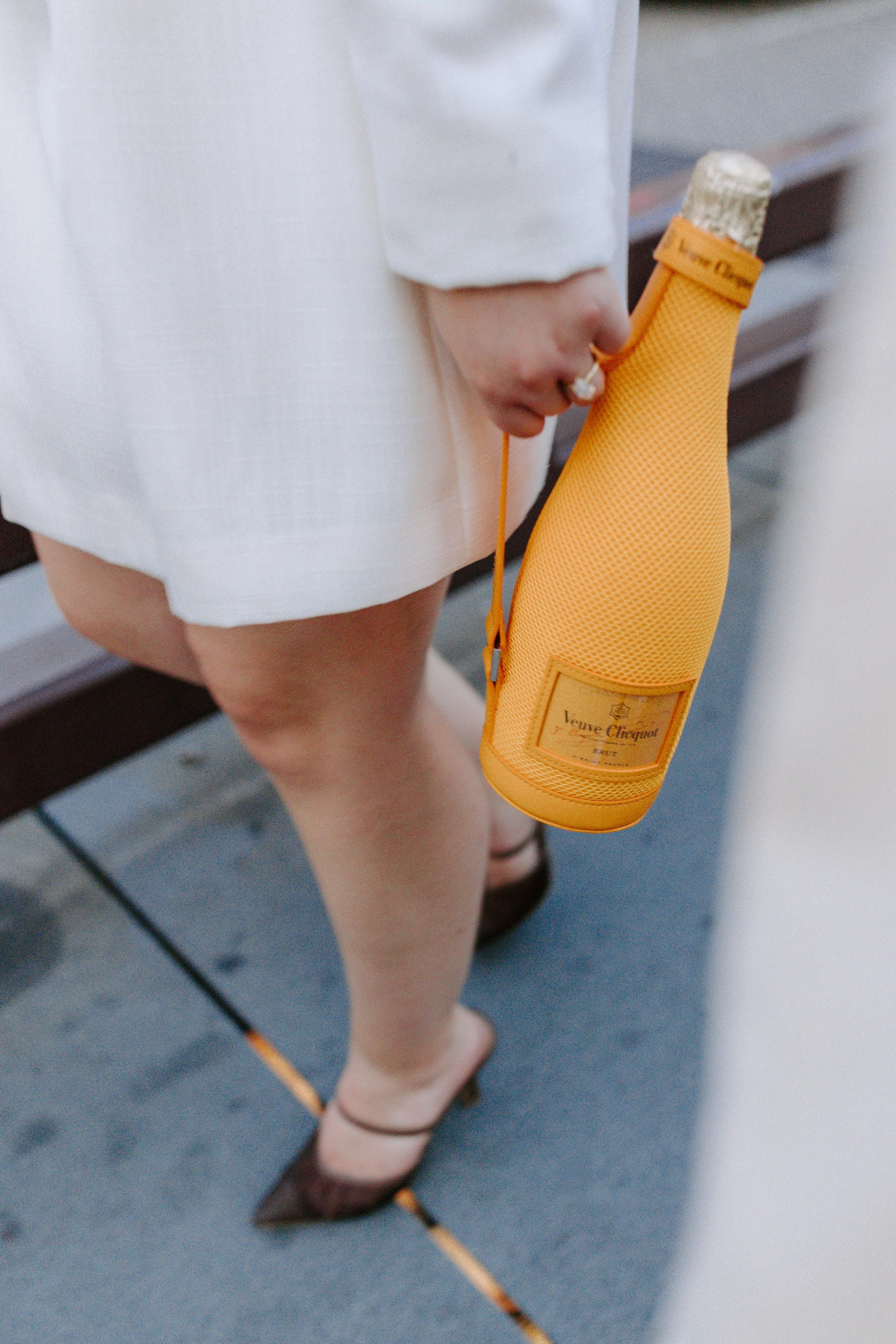 A person holding a yellow Veuve Clicquot champagne bottle in a protective cover, dressed in a white outfit, with brown high heels, standing on a gray surface.