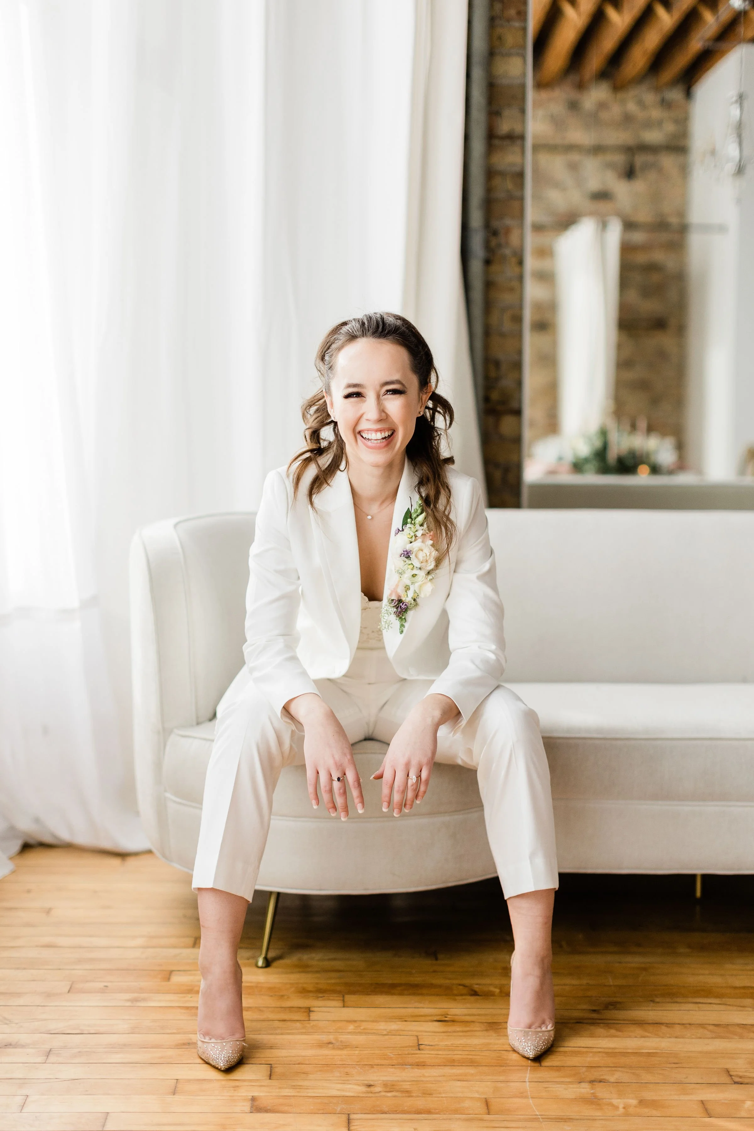 A woman dressed in a white suit, smiling and sitting on a cream-colored sofa in a bright room with wooden floors and exposed brick walls.