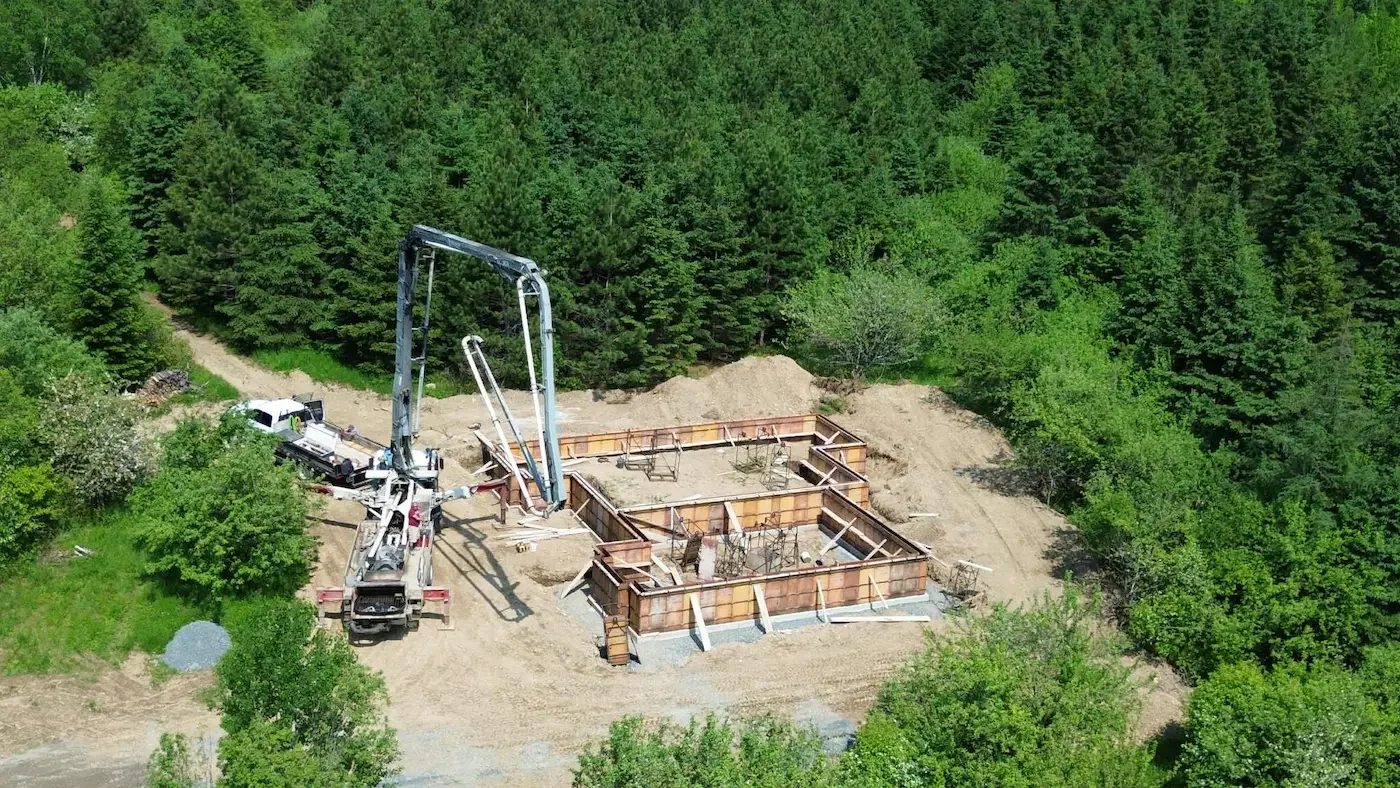 Aerial view of a construction site in a forested area, showing foundation walls, construction equipment, and a concrete pump truck.