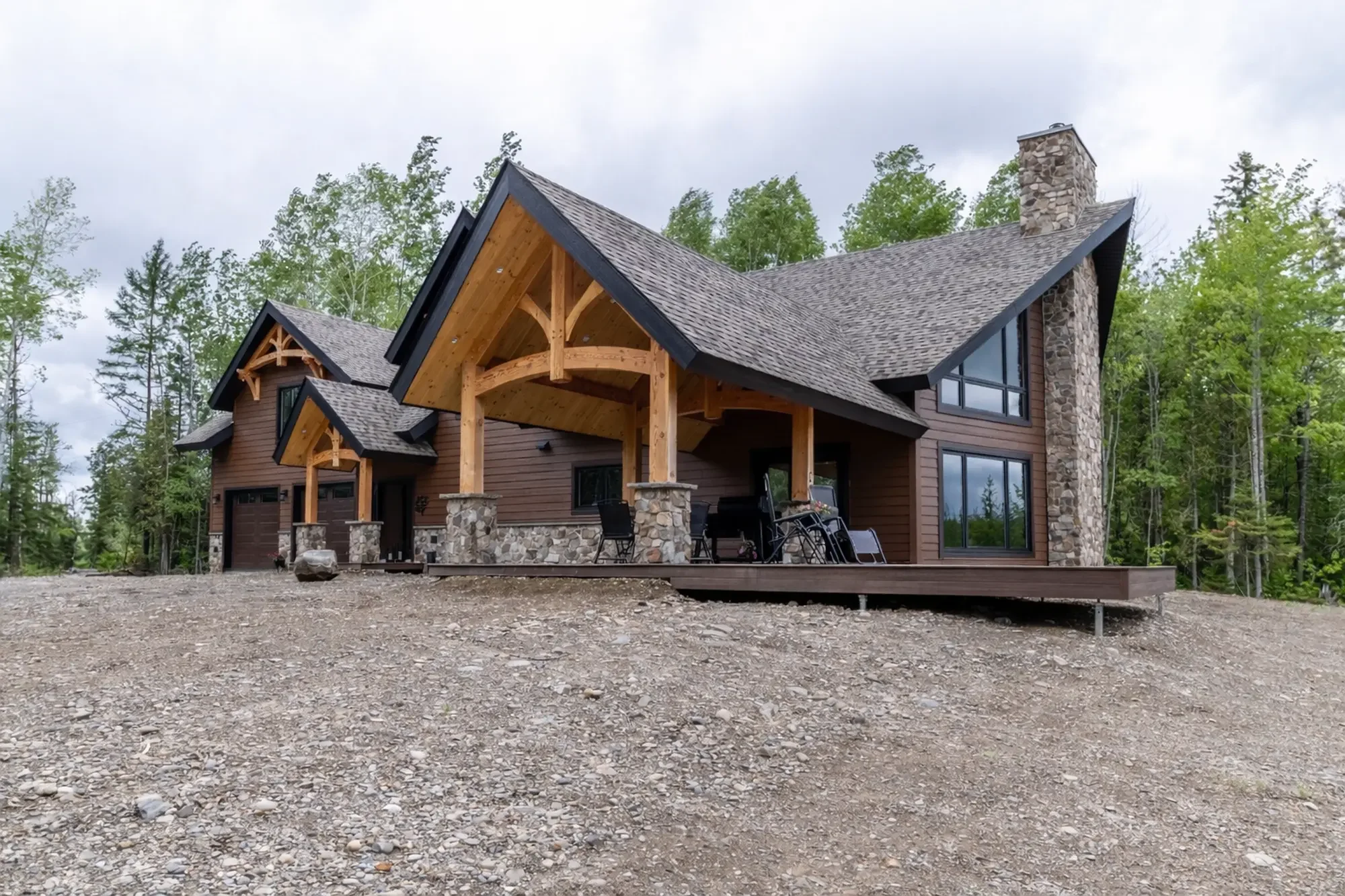 A modern house with a steeply pitched roof, stone chimney, large windows, and a wooden porch with supporting logs, located in a wooded area.