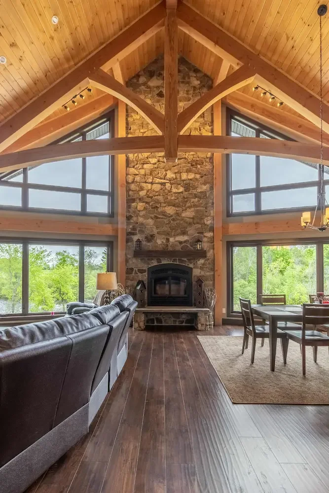 Living room with large windows, stone fireplace, wooden ceiling beams, and hardwood floors.
