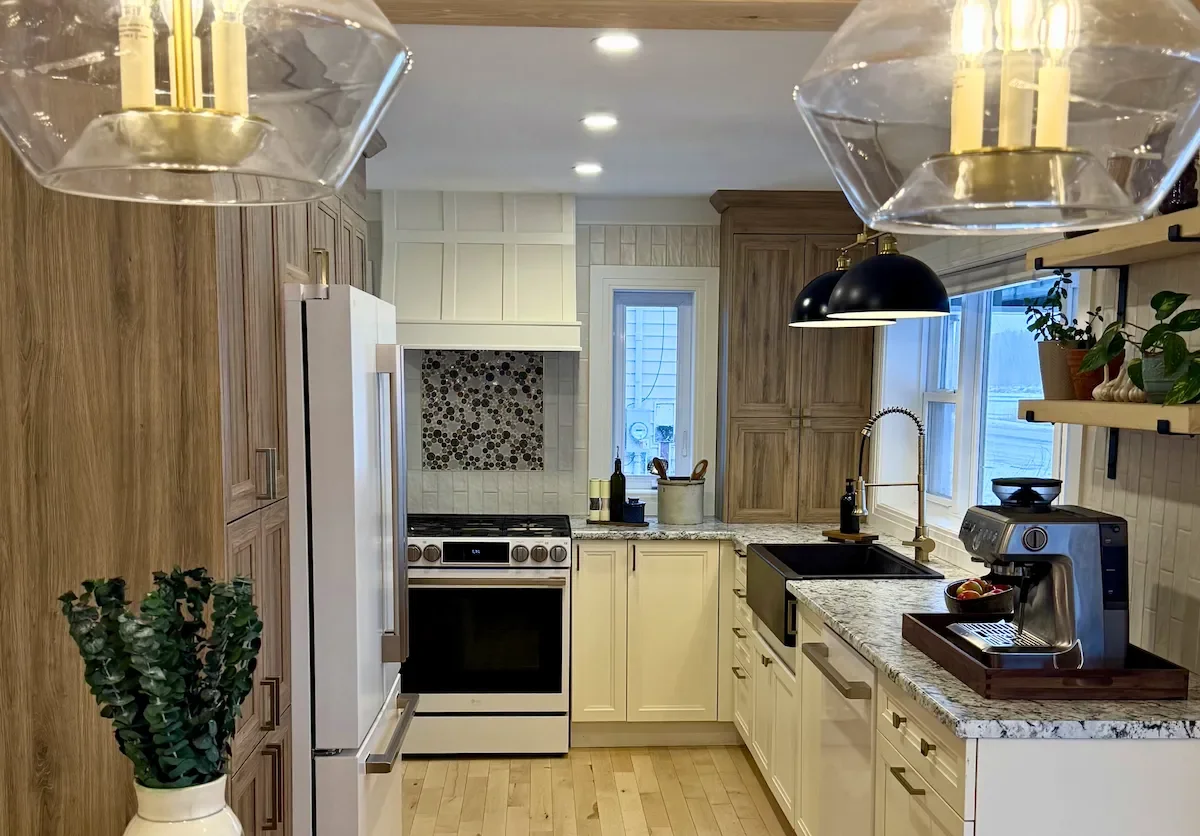 Modern kitchen with wooden cabinets, granite countertops, a black farmhouse sink, and a coffee maker. Ceiling lights and natural light through windows illuminate the space, with plants and kitchen utensils present.