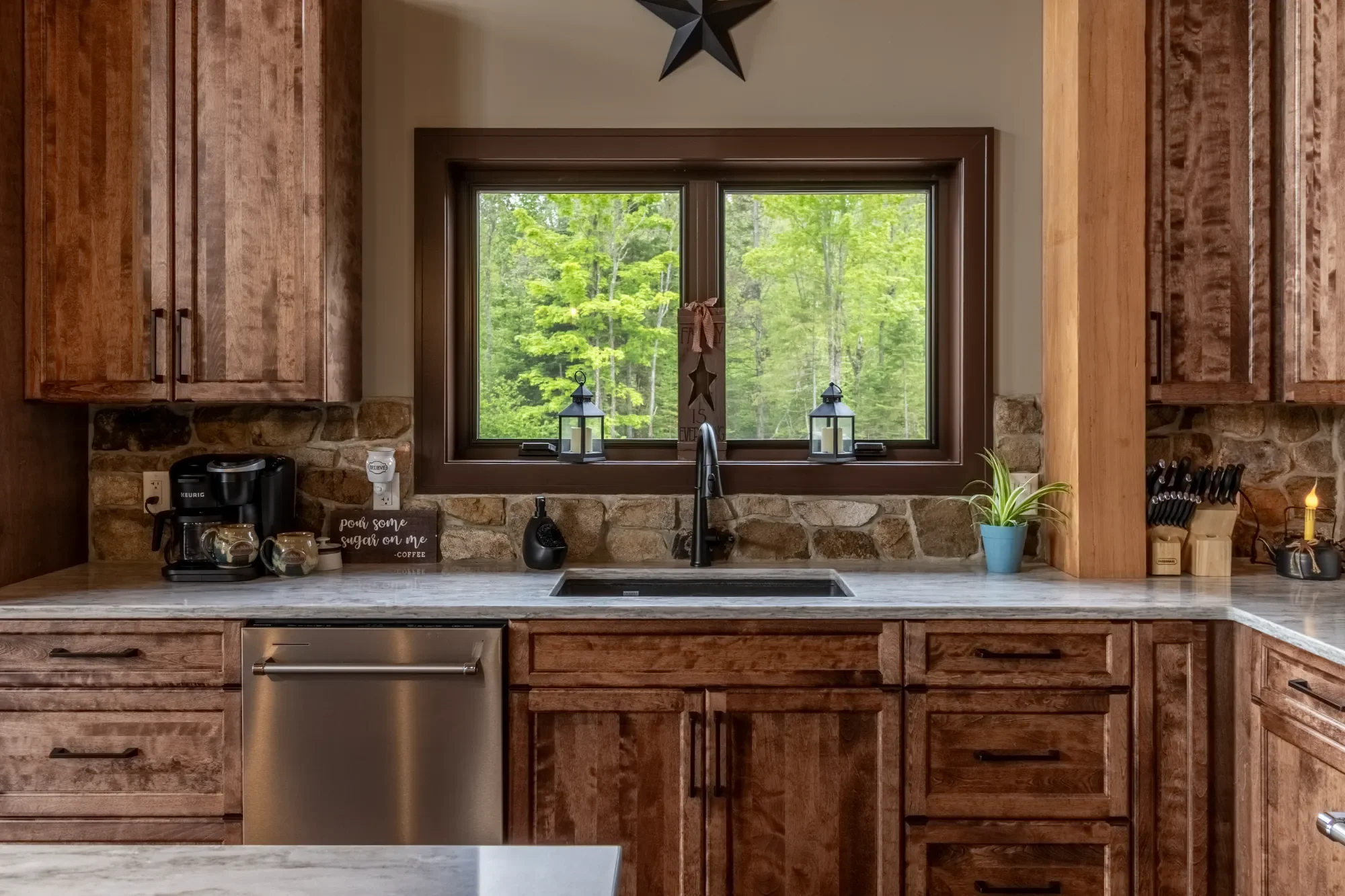 Kitchen with wooden cabinets, stone backsplash, a window with two lanterns on the windowsill, a coffee maker, potted plant, and kitchen utensils on the countertop.