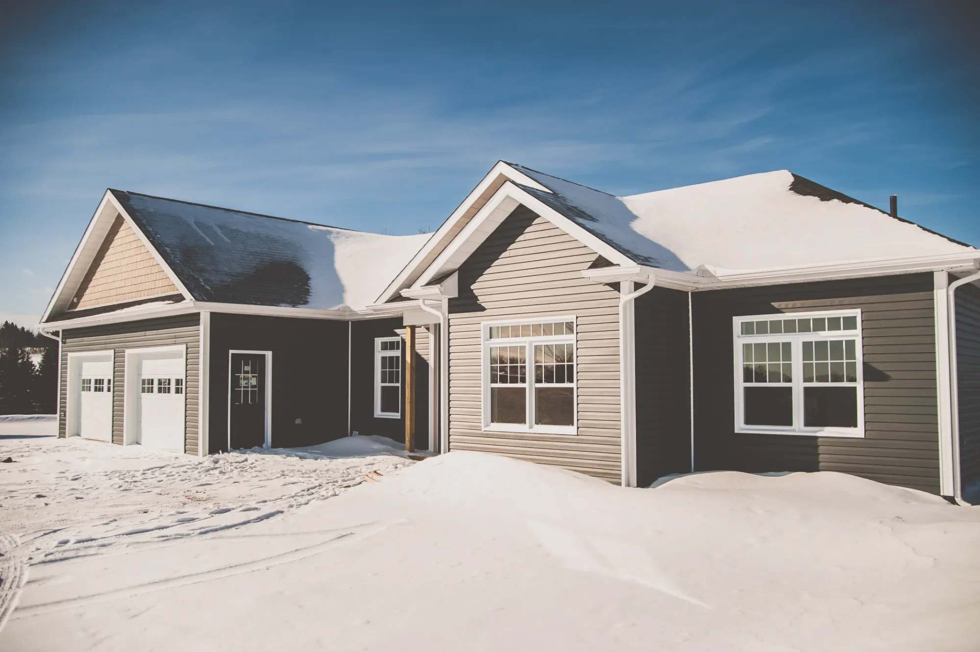 A modern house with gray siding, white trim, and snow-covered roof, surrounded by snow on the ground on a clear winter day.