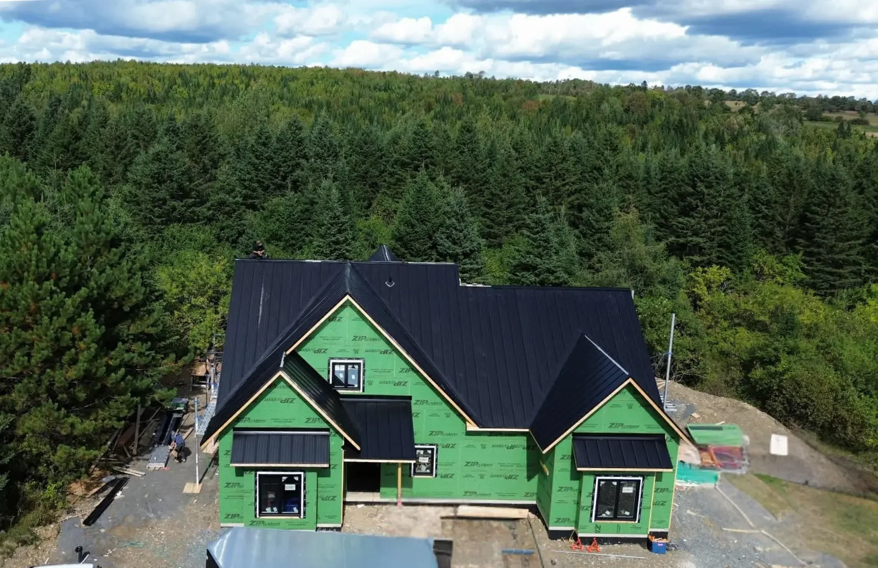 A house under construction with a black metal roof, green sheathing, surrounded by trees and a partly cloudy sky.