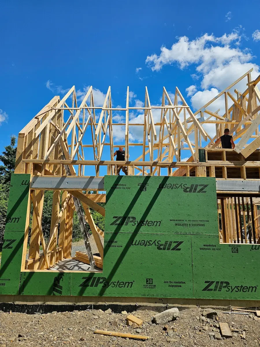 Construction site of a wooden house with framing in progress; two workers on the upper level, green ZIP system sheathing on the lower walls, blue sky with clouds in the background.