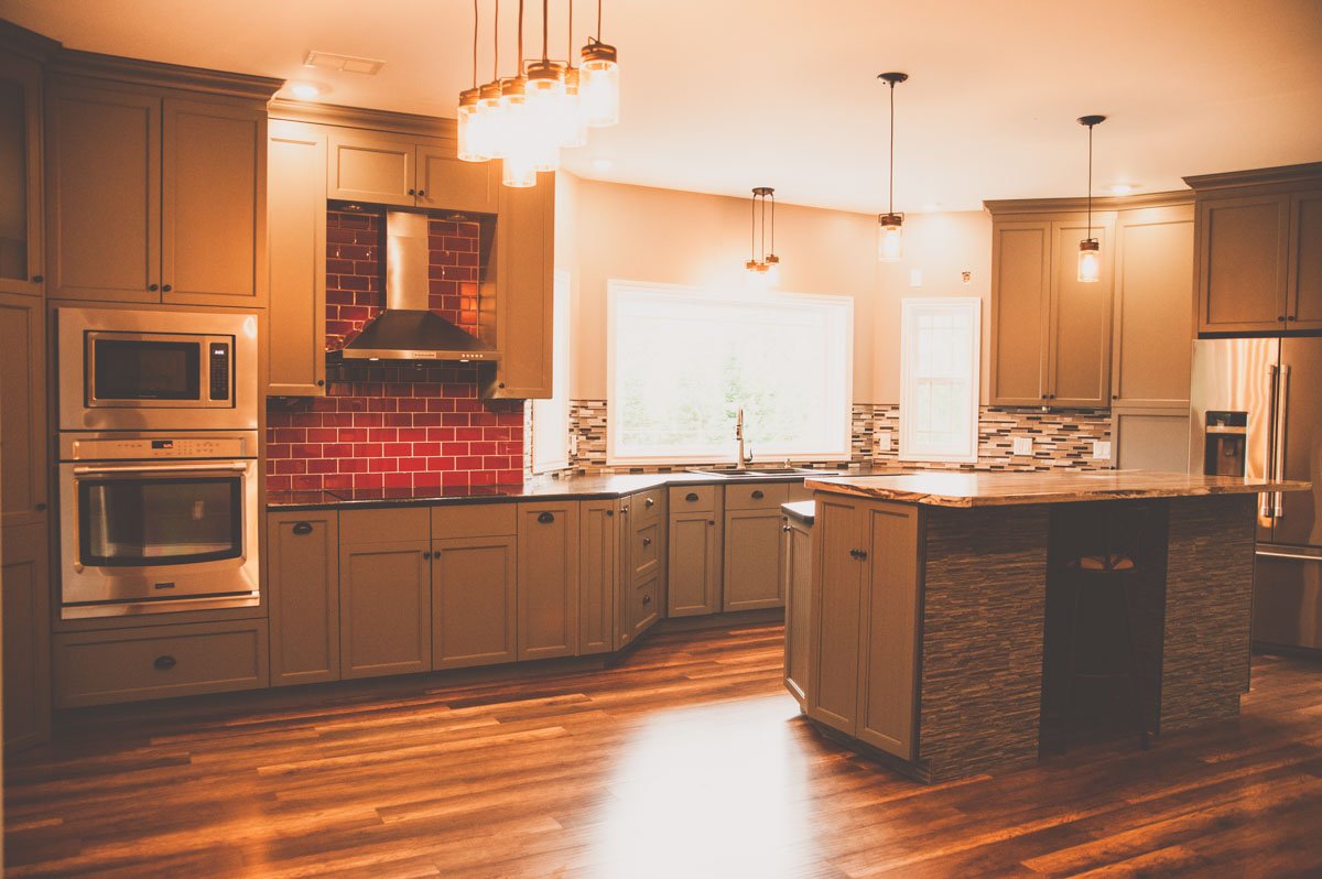 Modern kitchen with beige cabinets, red brick backsplash behind the stove, stainless steel appliances, wooden flooring, and a central island with a stone facade, illuminated by pendant lights.