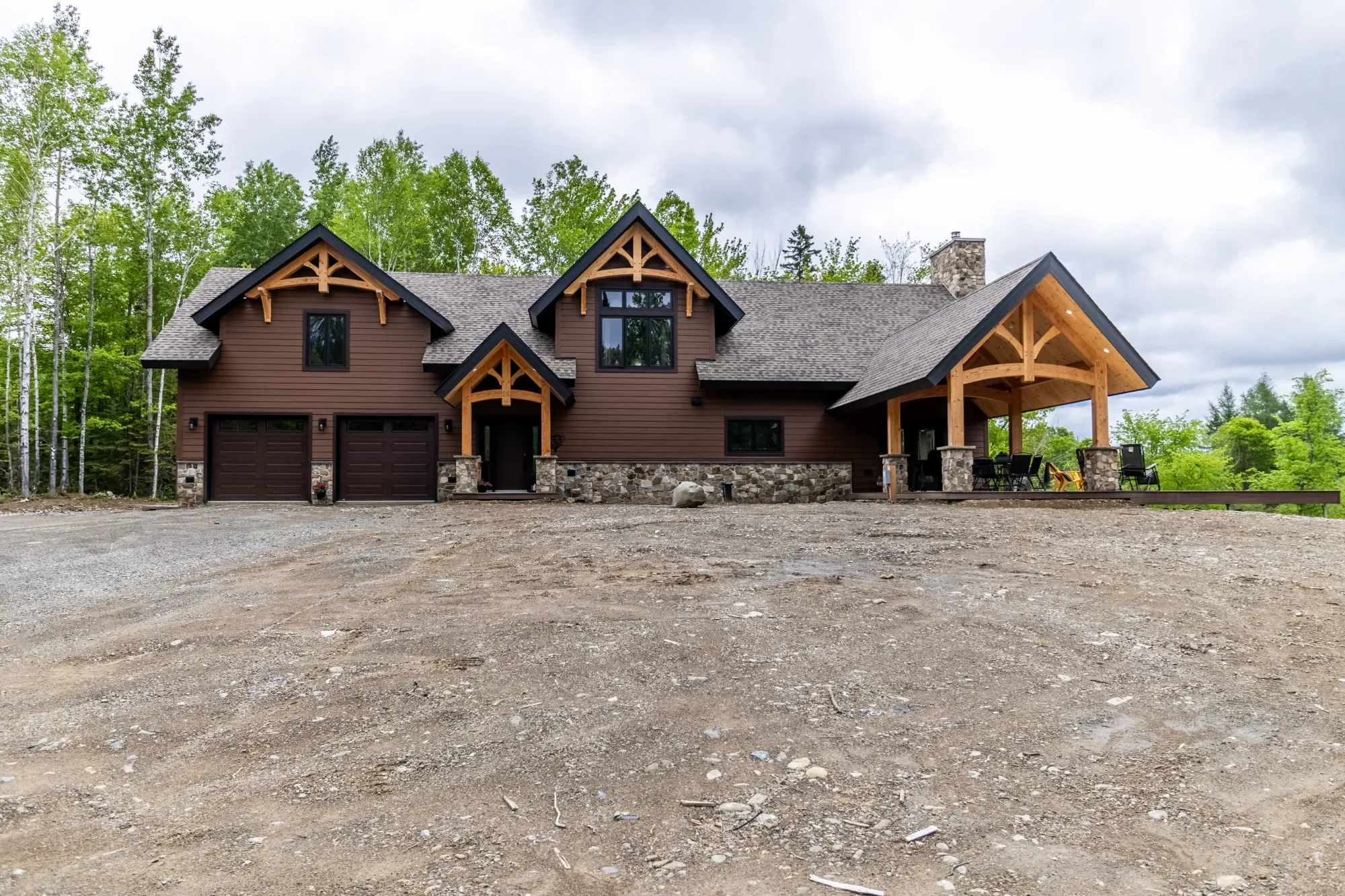Modern house with a brown exterior, stone accents at the base, and wooden trims, situated in a rural area with trees in the background and a large, unpaved driveway in front.