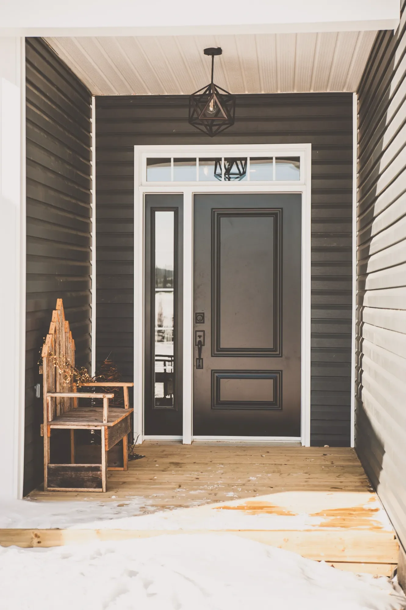 Front porch of a house with a black door, sidelights, transom window, and a geometric pendant light fixture. There is a wooden bench on the left side and snow on the porch floor.