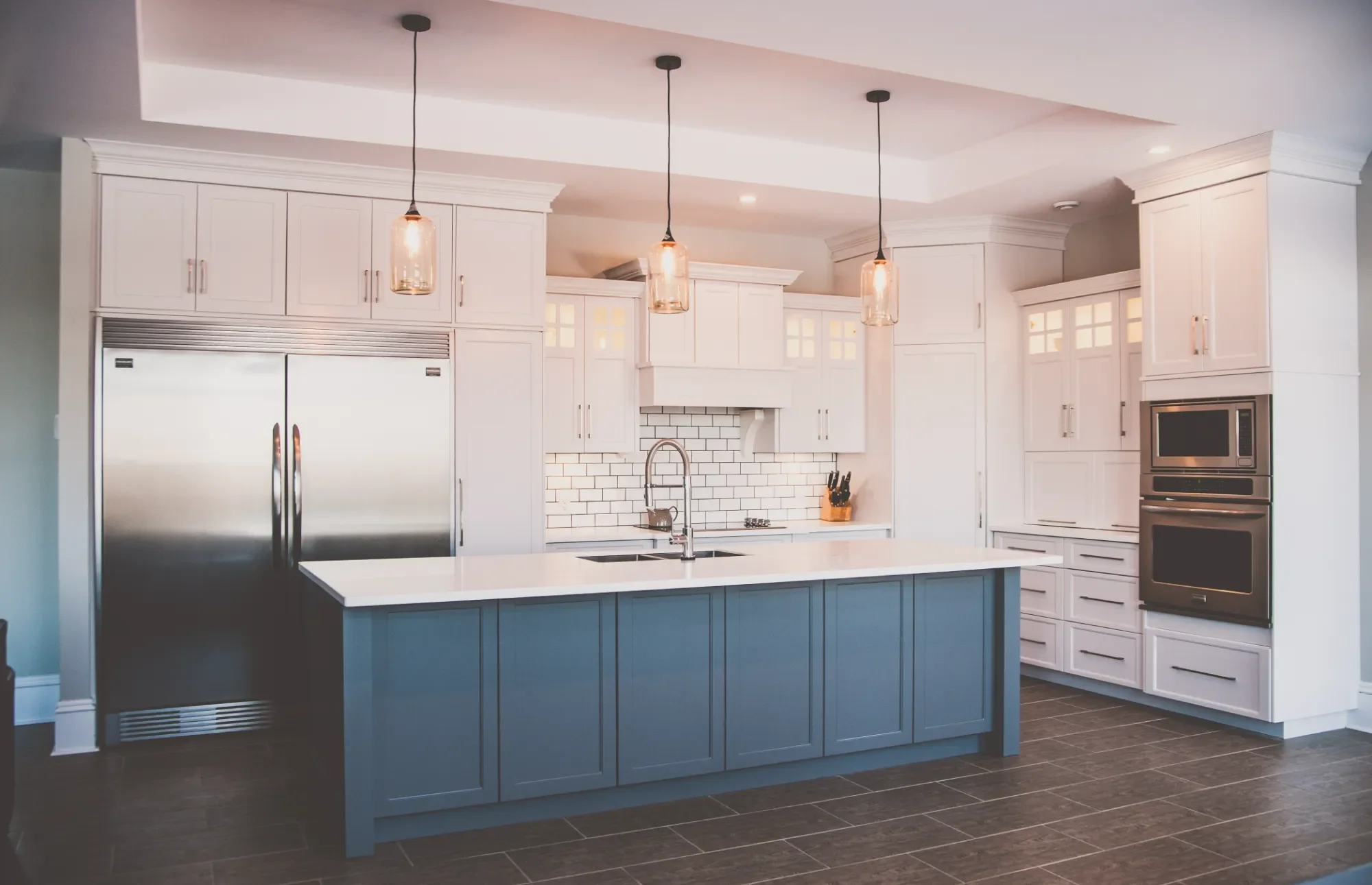 Modern kitchen with white cabinetry, a blue island with a white countertop, stainless steel refrigerator, built-in oven, and subway tile backsplash, illuminated by pendant lights.