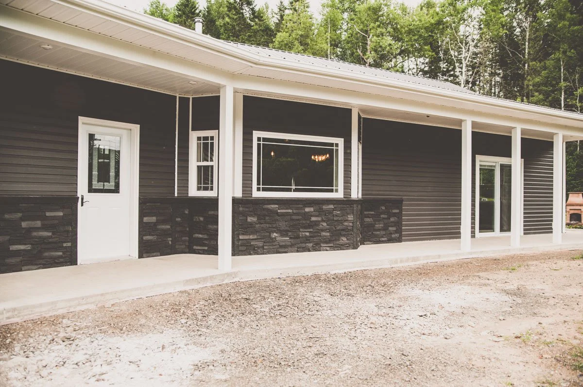 A modern house with gray siding, stone accents, a white door, and large windows, surrounded by trees and a gravel driveway.