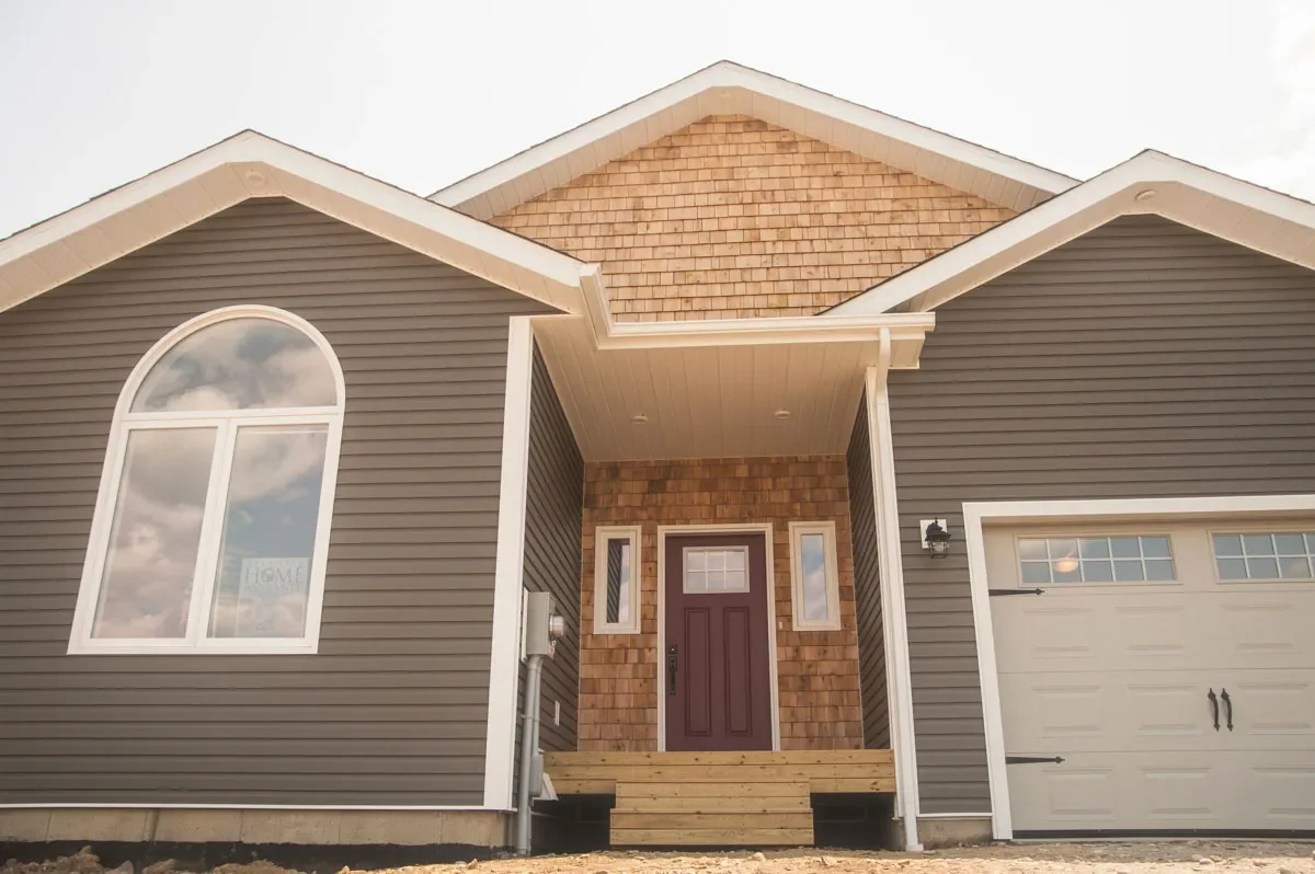 Newly constructed gray house with white trim and small wooden stairs leading to the front door, with a lawn and trees in the background.