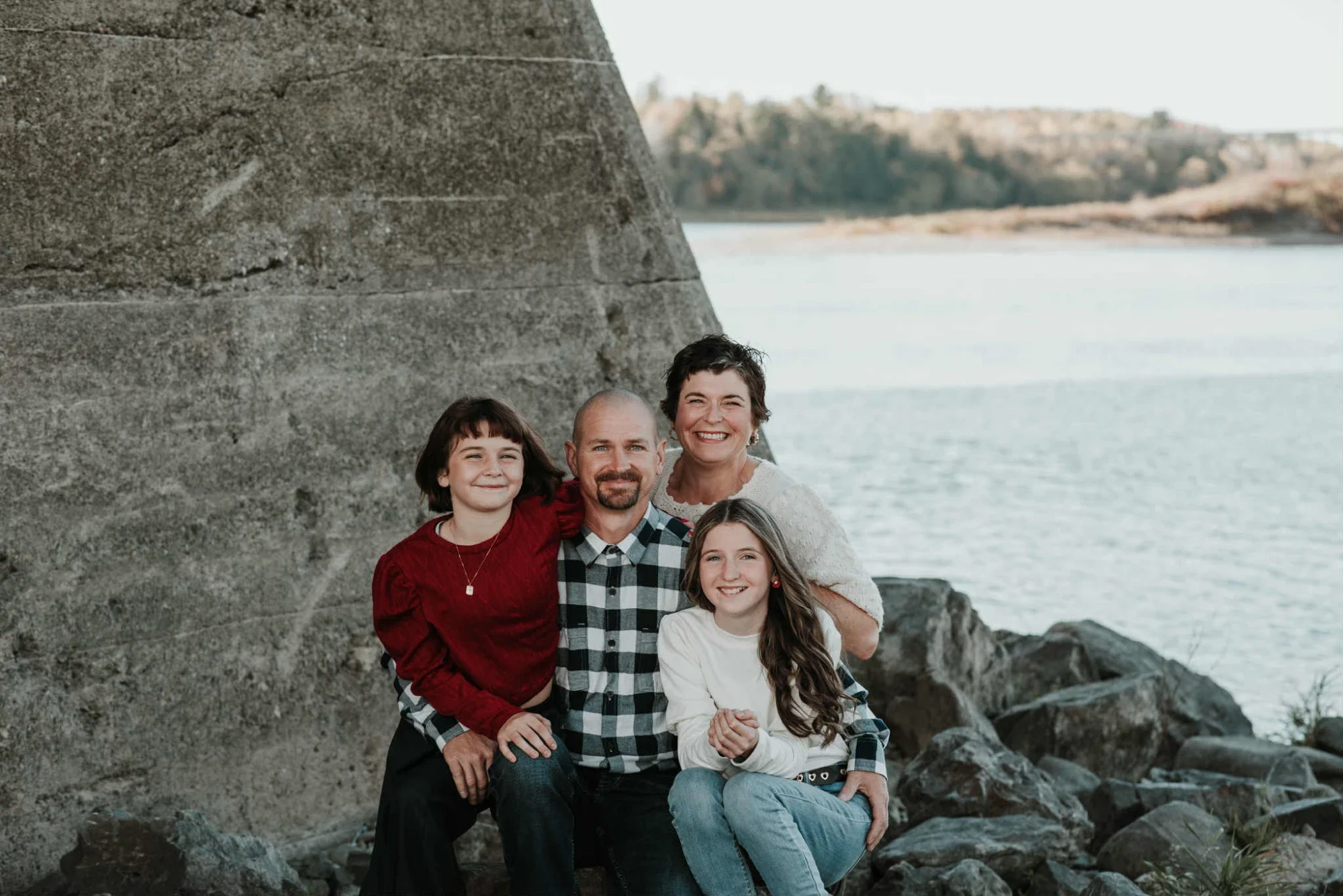 A family of four standing or sitting on rocks near a body of water with a large stone structure behind them, smiling for the camera.