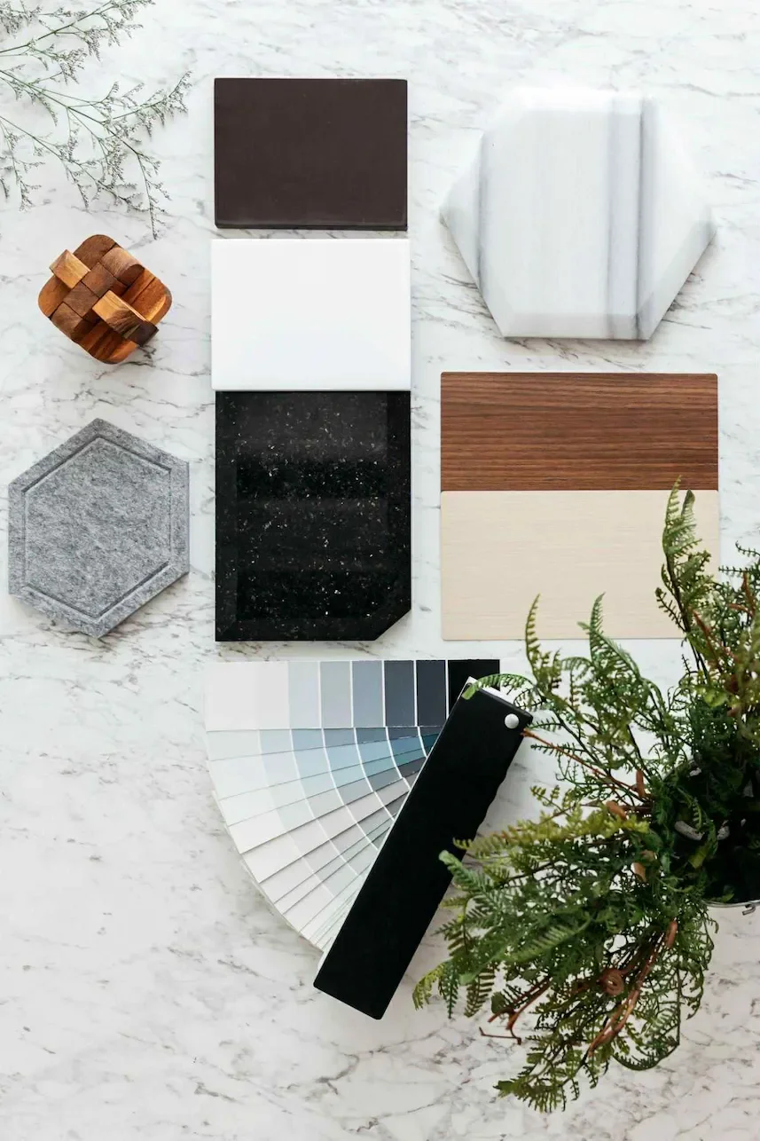 An assortment of interior design materials and samples on a marble surface, including paint swatches, wood and marble tiles, fabric or wallpaper samples, a clump of greenery, and a decorative wooden object.