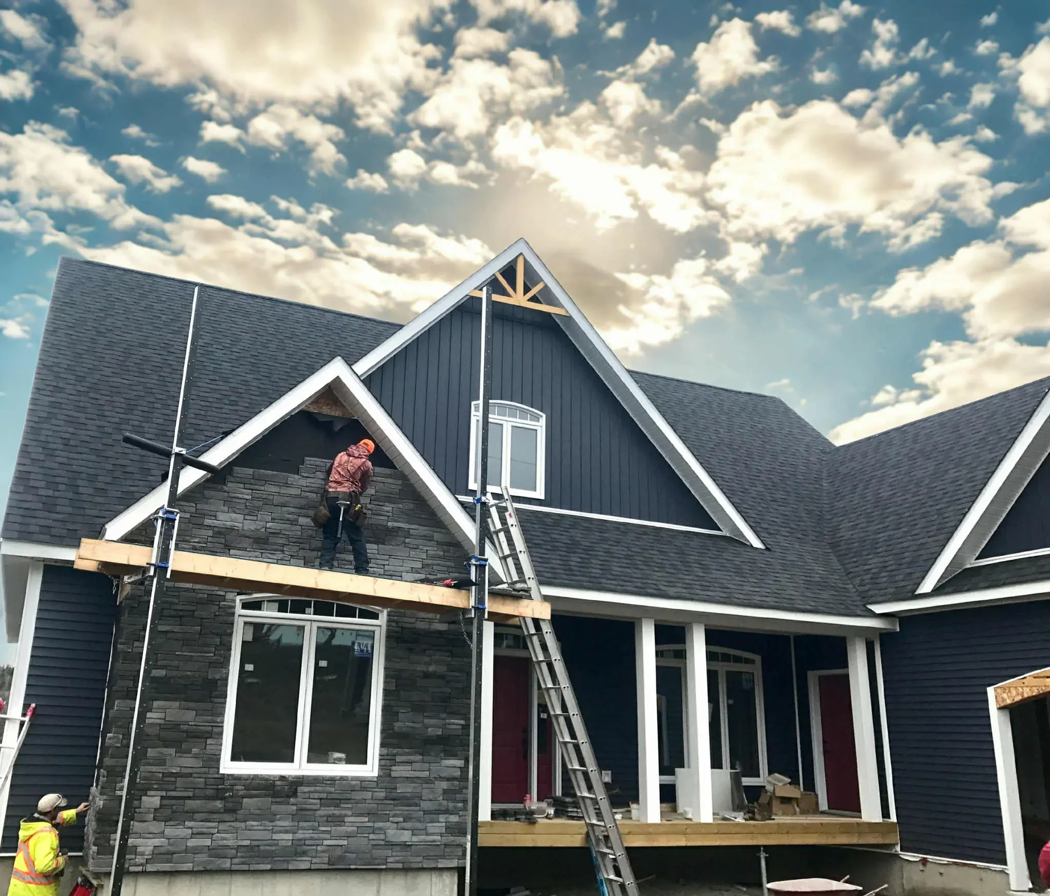 Construction workers are installing gray stone siding on a house with black siding and a sloped roof. One worker is on a scaffold, another on the ground.