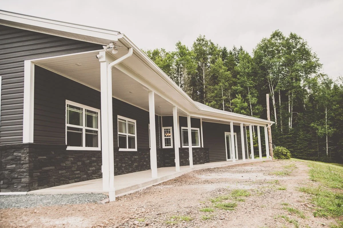 A house with dark siding, white window frames, and a white porch with multiple columns, set against a backdrop of trees and a cloudy sky.