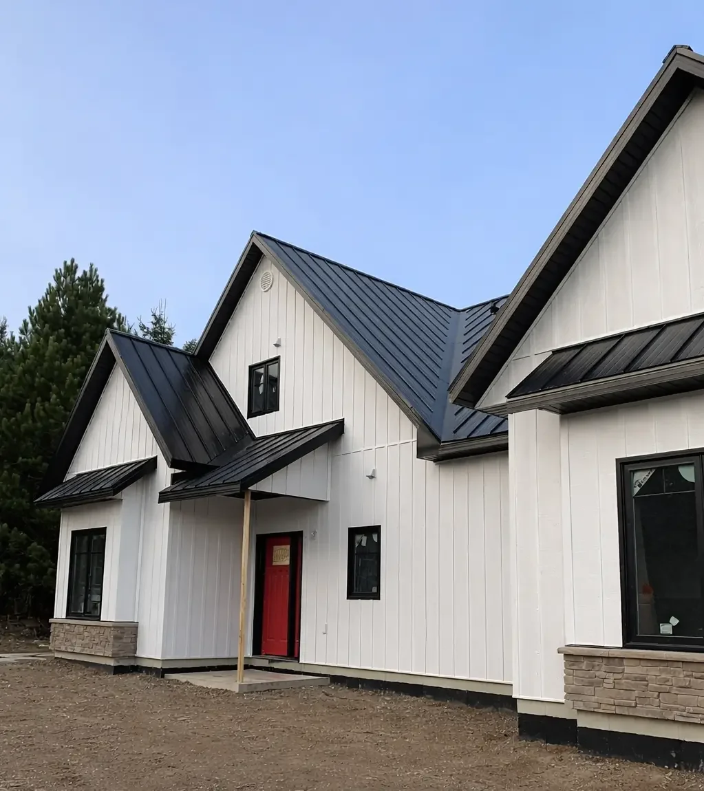 A house under construction with a black metal roof, located in a wooded area with dense trees and a hilly background.