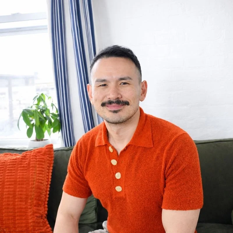 This smiling Asian American man with a mustache is a LCSW psychotherapist appearing in his private office in Brooklyn, NY, where he treats issues like grief, life transitions, depression, anxiety, trauma, LGBTQ identity, and men's issues.