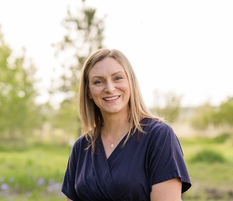 A smiling woman with light brown hair in a navy blue top outdoors with trees and grass in the background
