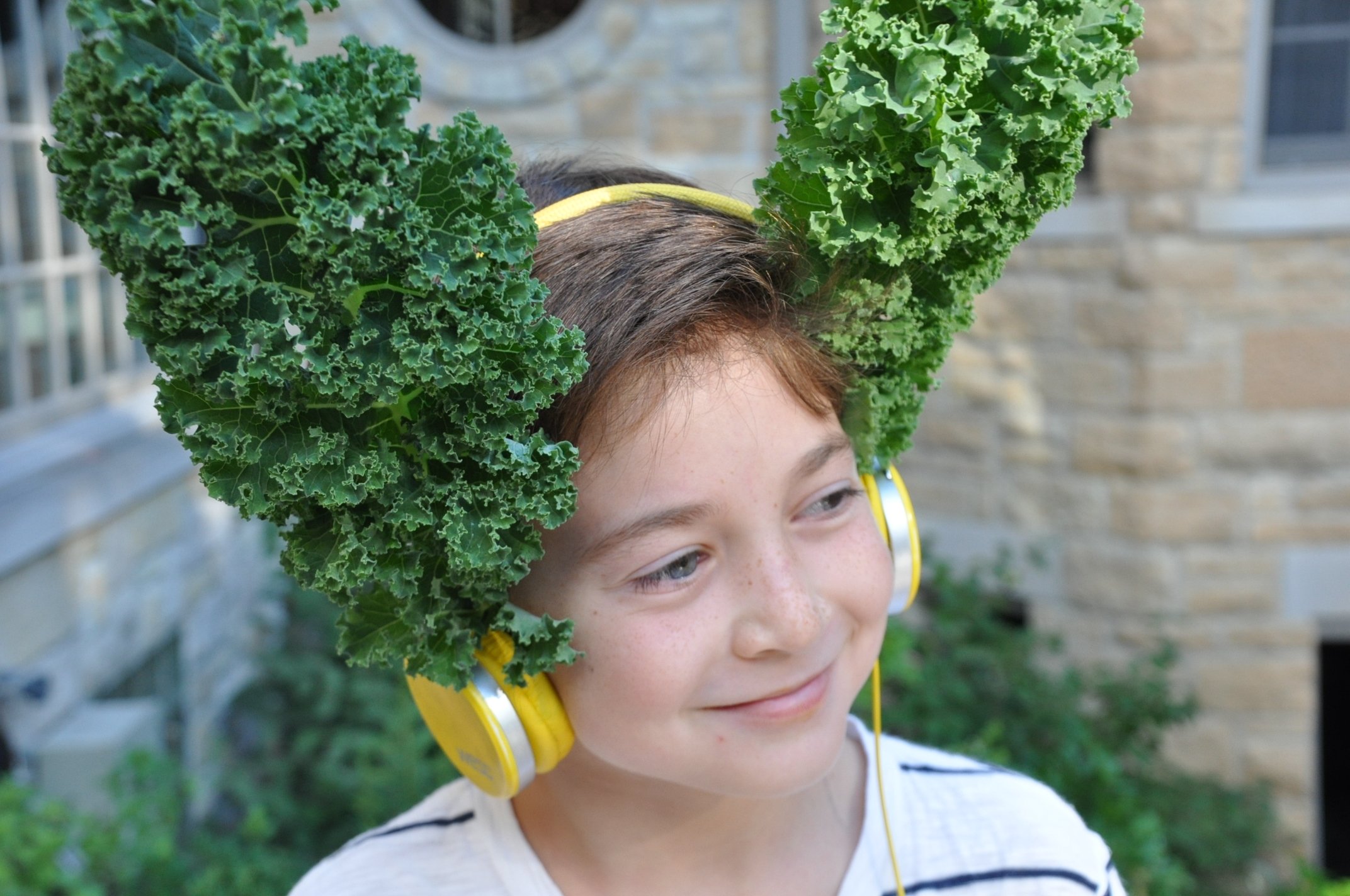 A young boy with curly brown hair and freckles wearing yellow headphones and a white shirt with black stripes. He has kale leaves on his head and is smiling, outdoors in front of a brick building.