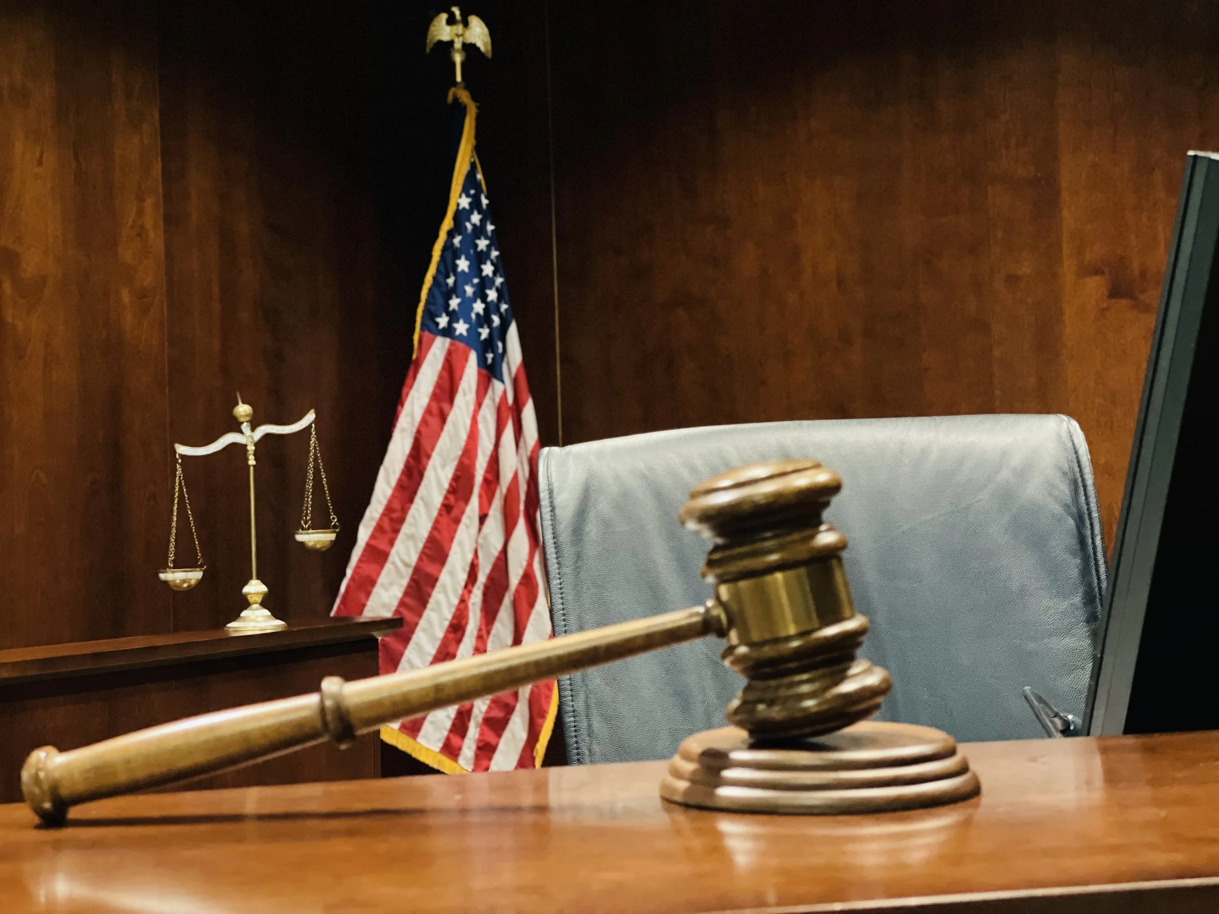 A judge's gavel on a wooden desk, with a courtroom in the background including a chair, the United States flag, and a set of scales of justice.