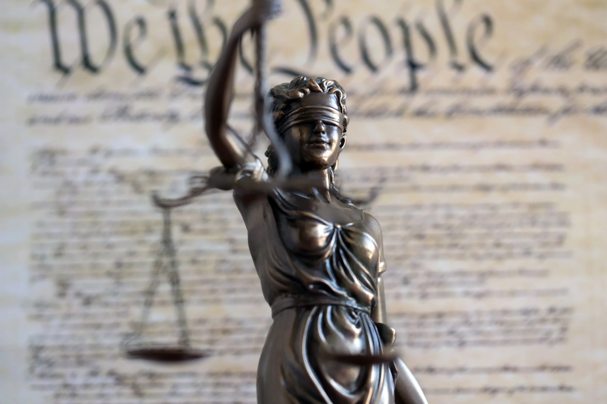 Close-up of a bronze statue of Lady Justice with a blindfold and raised sword, blurred historical document background.
