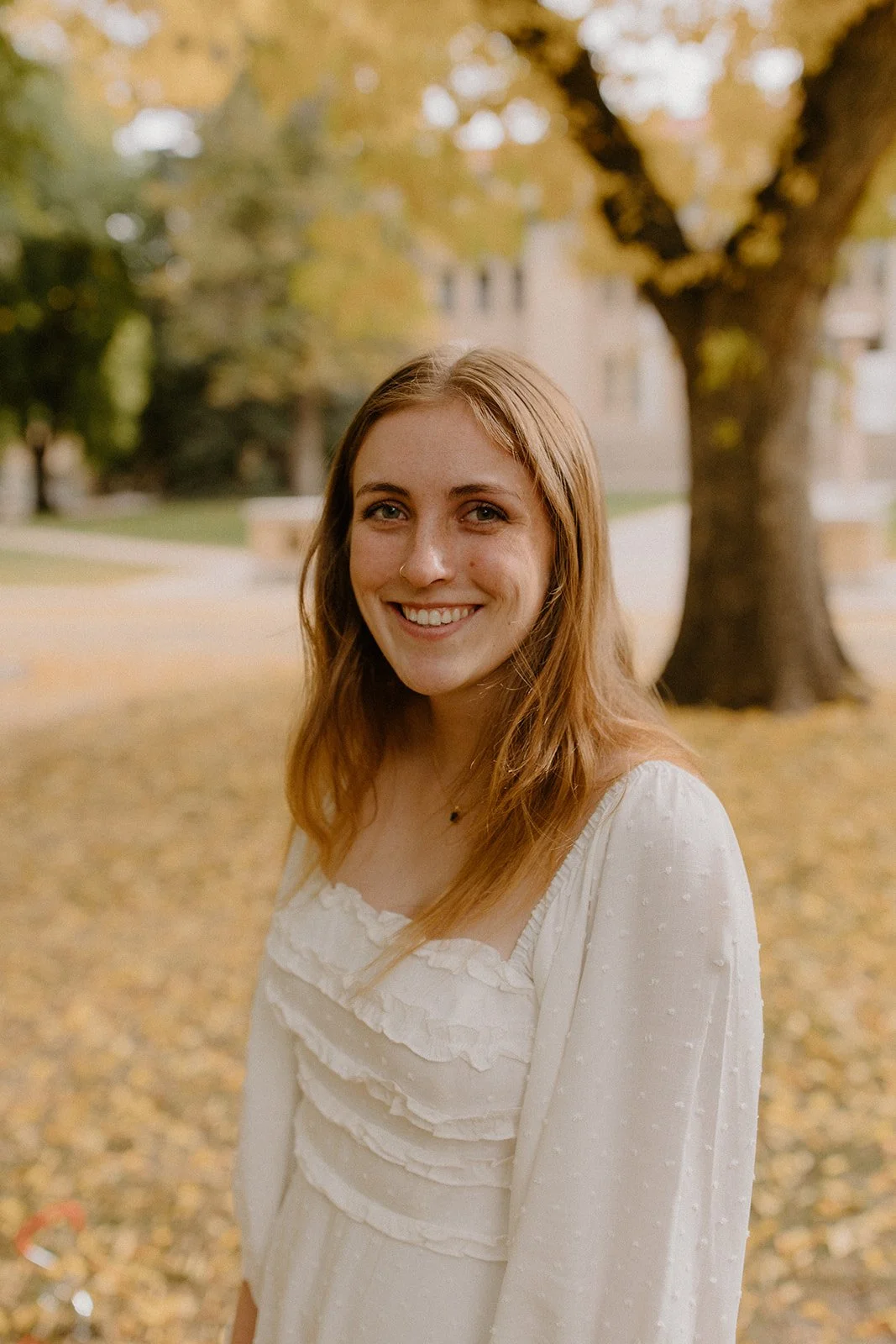 A young woman with red hair smiling outdoors in fall, standing in front of a leafless tree and yellow foliage.
