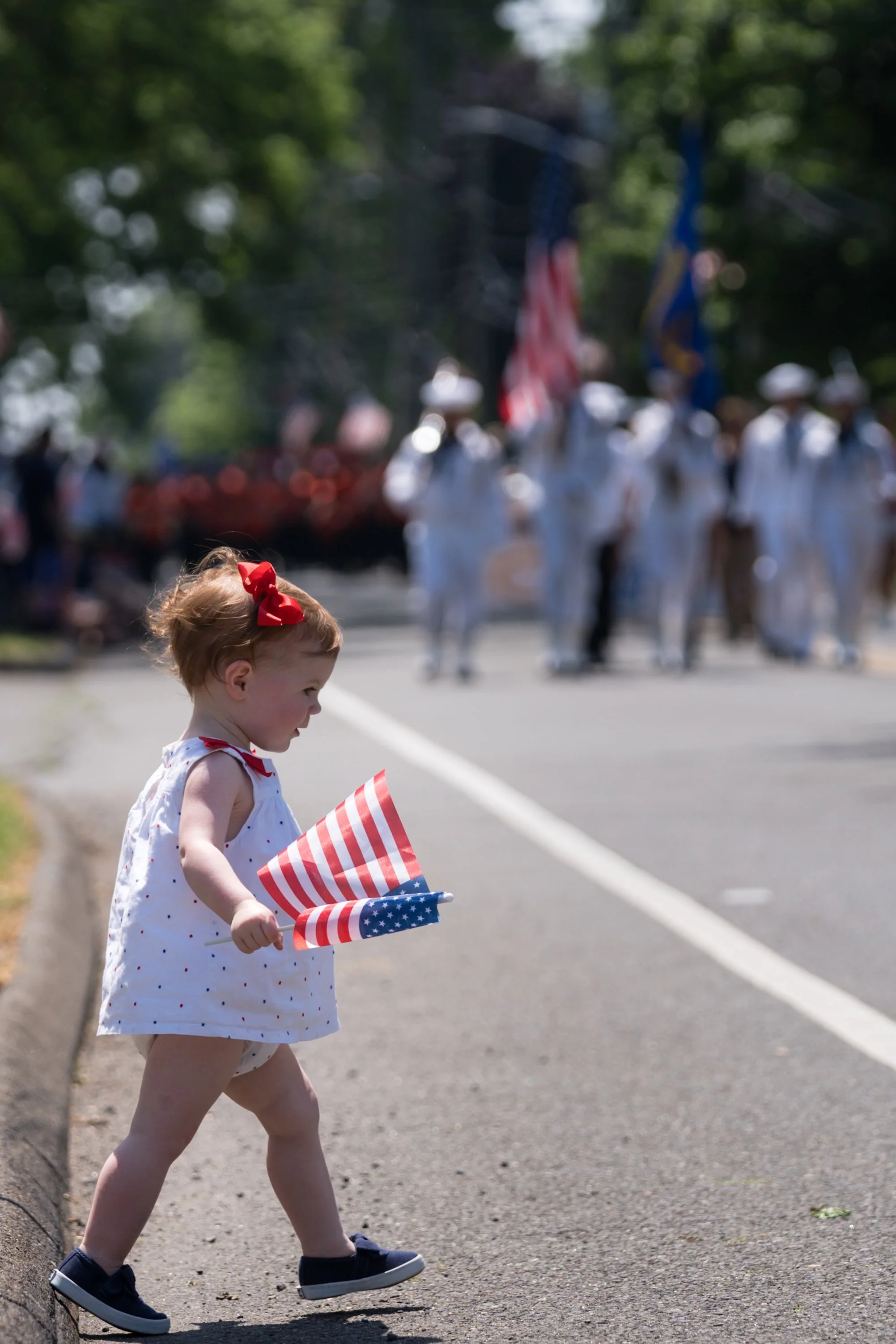 A young girl holding small American flags crossing the street during a parade, with a group of people marching in the background.