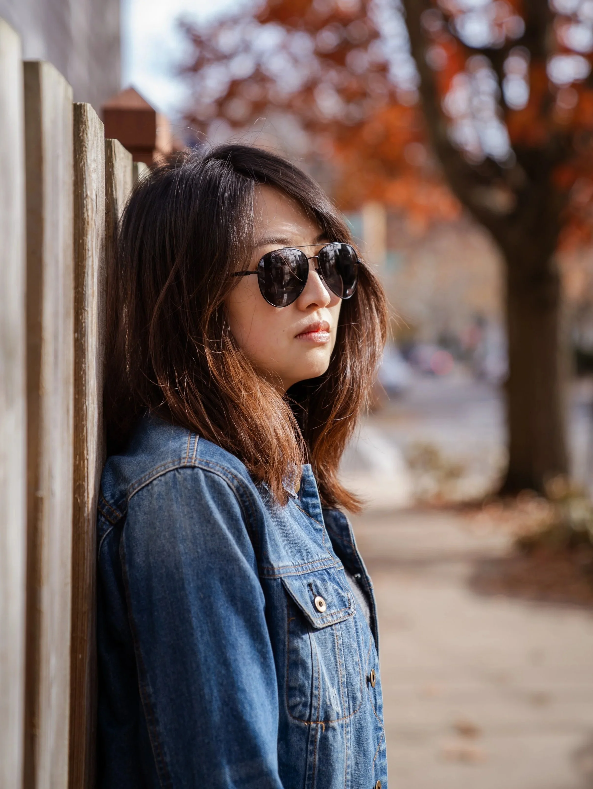 A woman with brown hair wearing sunglasses and a denim jacket leaning against a wooden fence. Behind her are autumn trees with orange leaves and a blurred street scene.