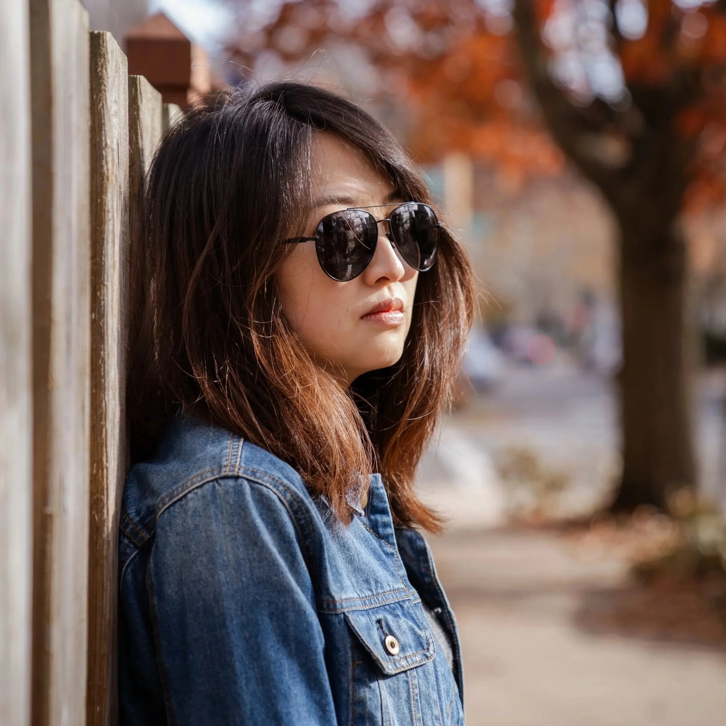 A young woman with brown hair wearing sunglasses and a denim jacket, leaning against a wooden fence outdoors during autumn.