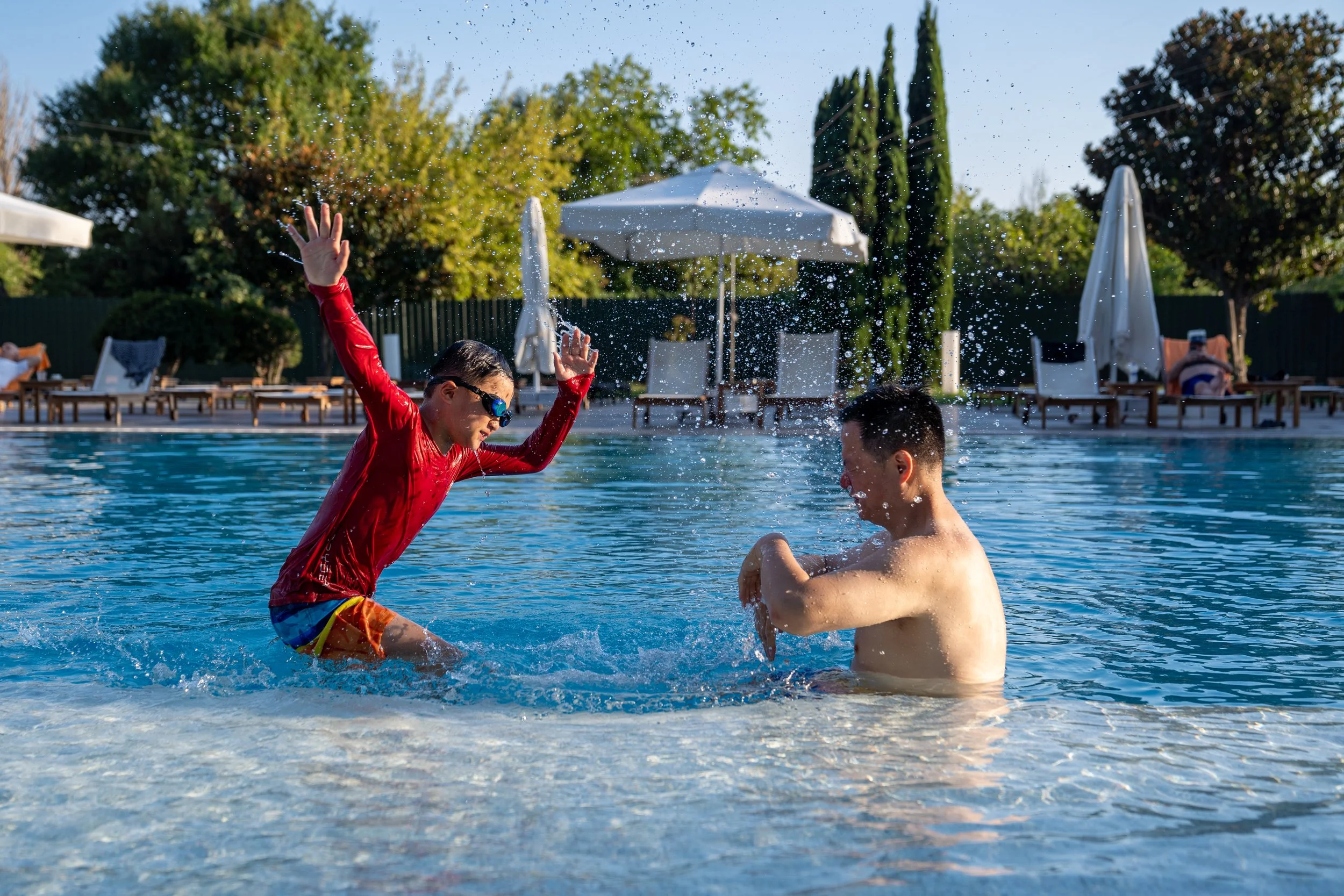 Two boys playing in a swimming pool, one wearing red shirt and sunglasses, the other shirtless. The boy in red is splashing water on the other boy, who is reacting with his head down, in a backyard pool setting with umbrellas and lounge chairs.